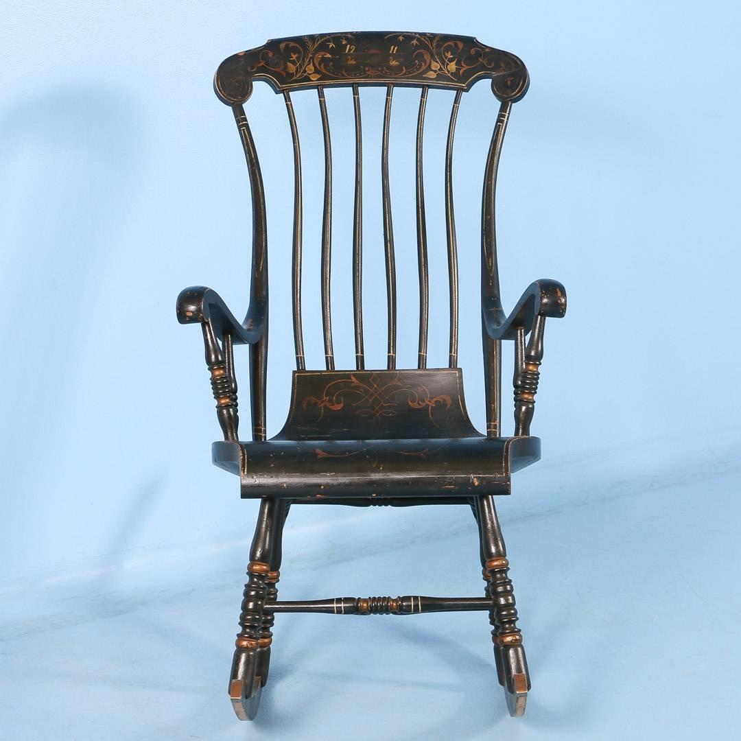 An antique spindle back rocker from Sweden dated 1911, with scrolled arms and supported by six legs and long sweeping rockers. The original painted details include a base color of black with accents of gold on the turned legs as well as elaborate