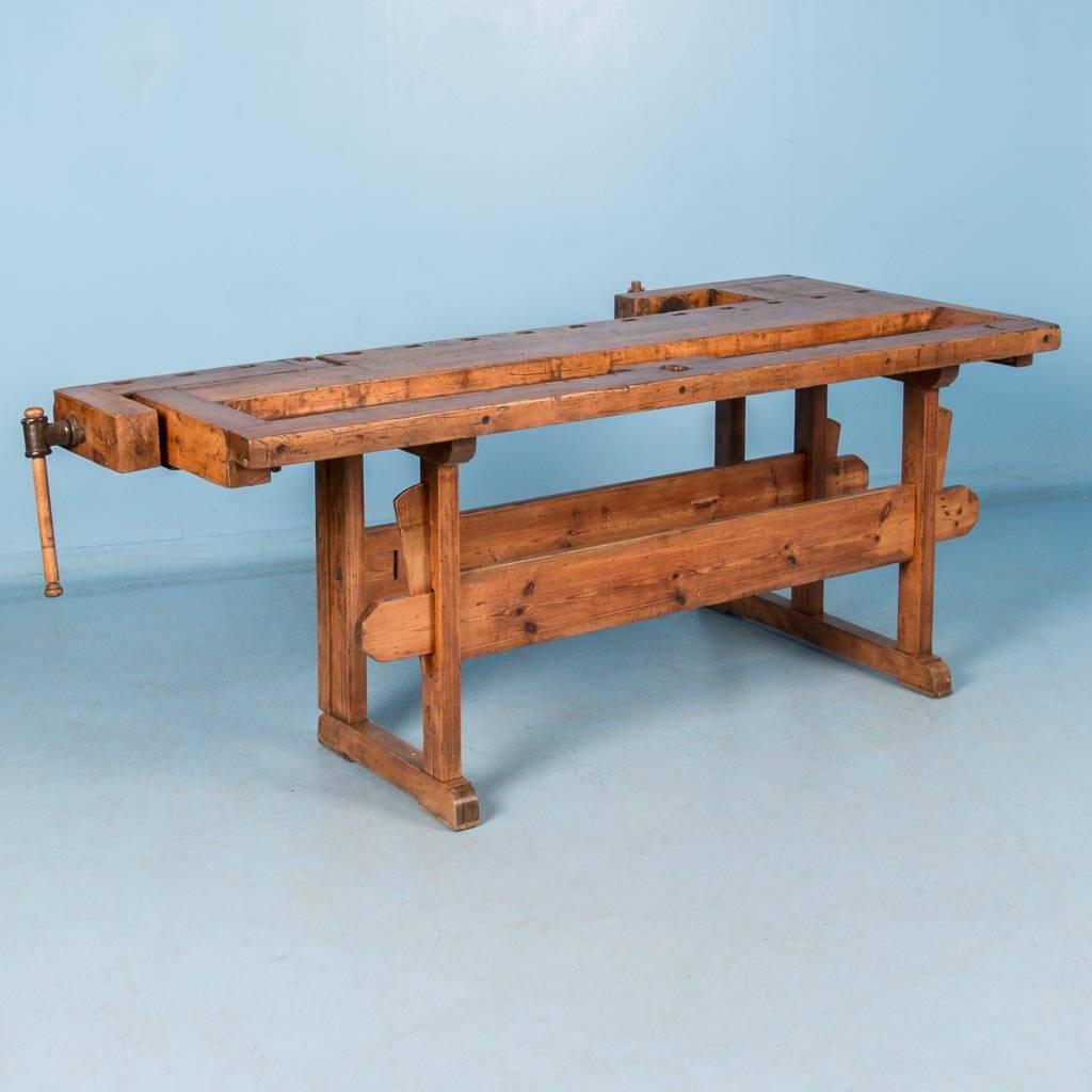 This antique hardwood workbench with a pine base features a pair of vices with wood and iron handles. The recessed tray is where the carpenter would lay his tools and was also used to catch debris. The traditional keyed trestle base allows it to be