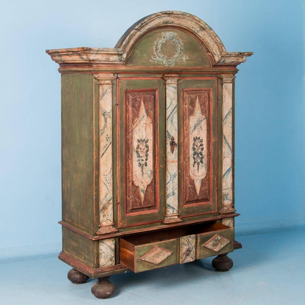 This mid-18th century baroque armoire features a molded arched pediment above paneled doors with a full drawer below, all resting on large bun feet. The original painted finish has red and cream colored accents over a green background. The armoire