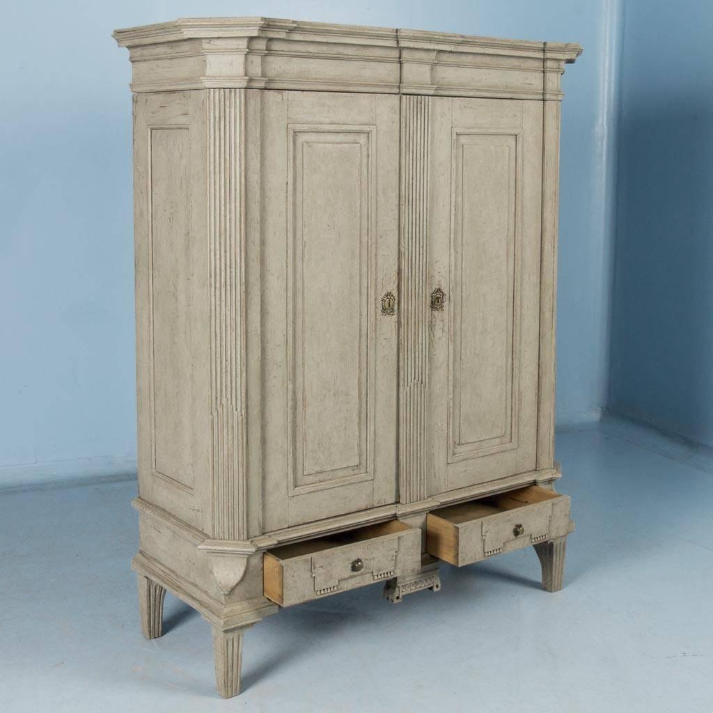 This early 19th century Swedish Gustavian armoire features a broad molded crown with mitered corners, paneled doors and a pair of small drawers below. The old painted finish appears gray at first glance, but on closer examination also appears a very