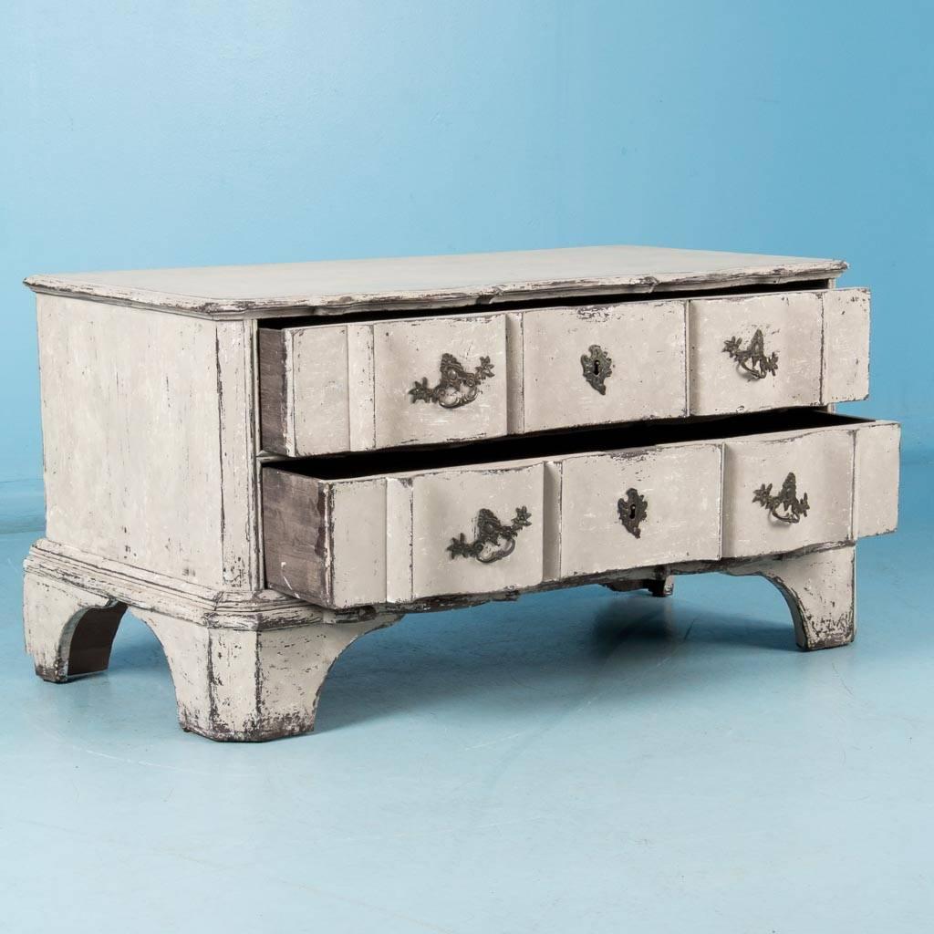 This unique mid-18th century low chest of drawers, circa 1760, has a molded and scalloped top which is carried down through the two drawer fronts resulting in an elegant, serpentine case. The natural pine shows through the distressed white paint.