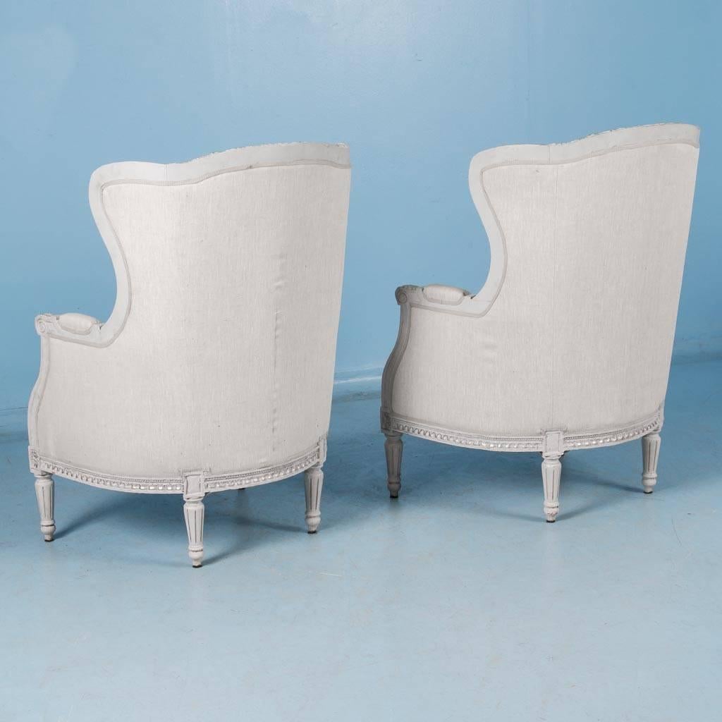 Pair of Swedish wingback chairs, circa 1920-1940, painted a light gray with silver highlights. Intricate floral carvings cover the high back frame which is covered in a natural ecru linen fabric. The chairs are clean, sturdy and comfortable and
