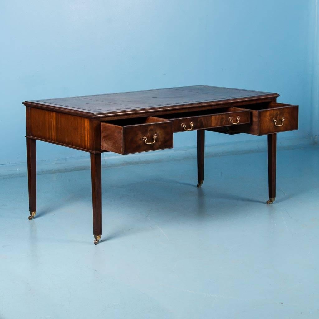 Antique mahogany desk from England with a hand tooled gold border on a brown leather top. The desk features three working drawers with brass hardware and tapered legs resting on original brass casters. Please take a moment to enlarge the photos and