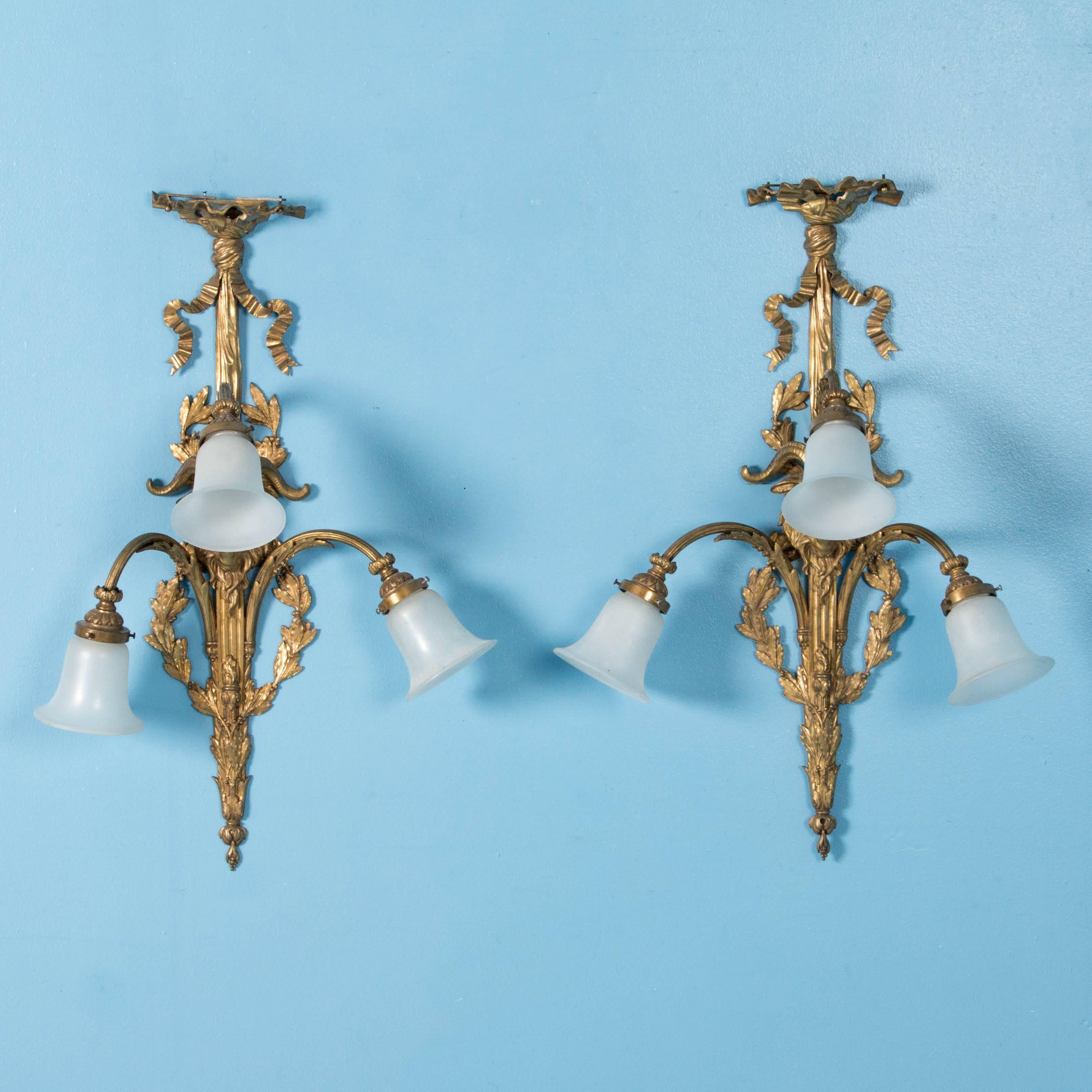 Each of the large ornate brass wall sconces has three goose neck arms with white frosted glass shades. The central ram's head is surrounded by a tied ribbon, wreath and leaf detail, all in brass. These are from a Victorian Blackhawk, Colorado home