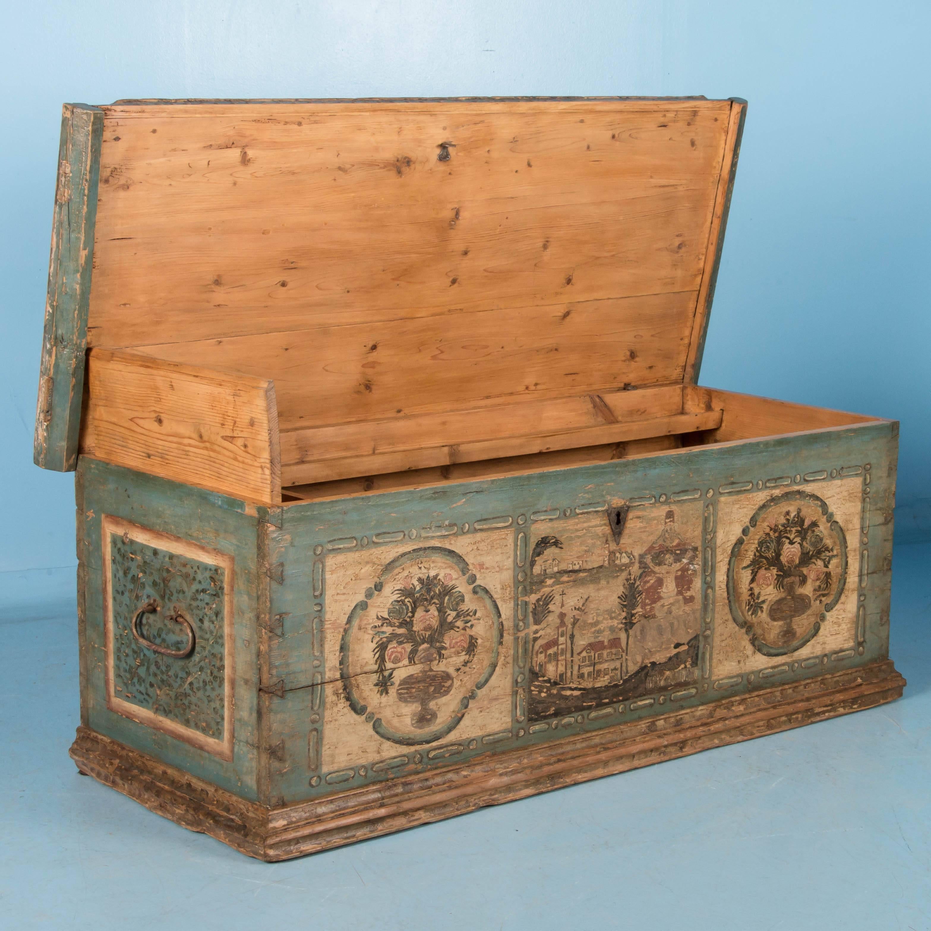 The original paint and details in this trunk are exceptional. The front of the trunk depicts a village scene and stylized vases of flowers in muted colors, set against a soft blue background. Inside is a shallow tray and a small utility box with a
