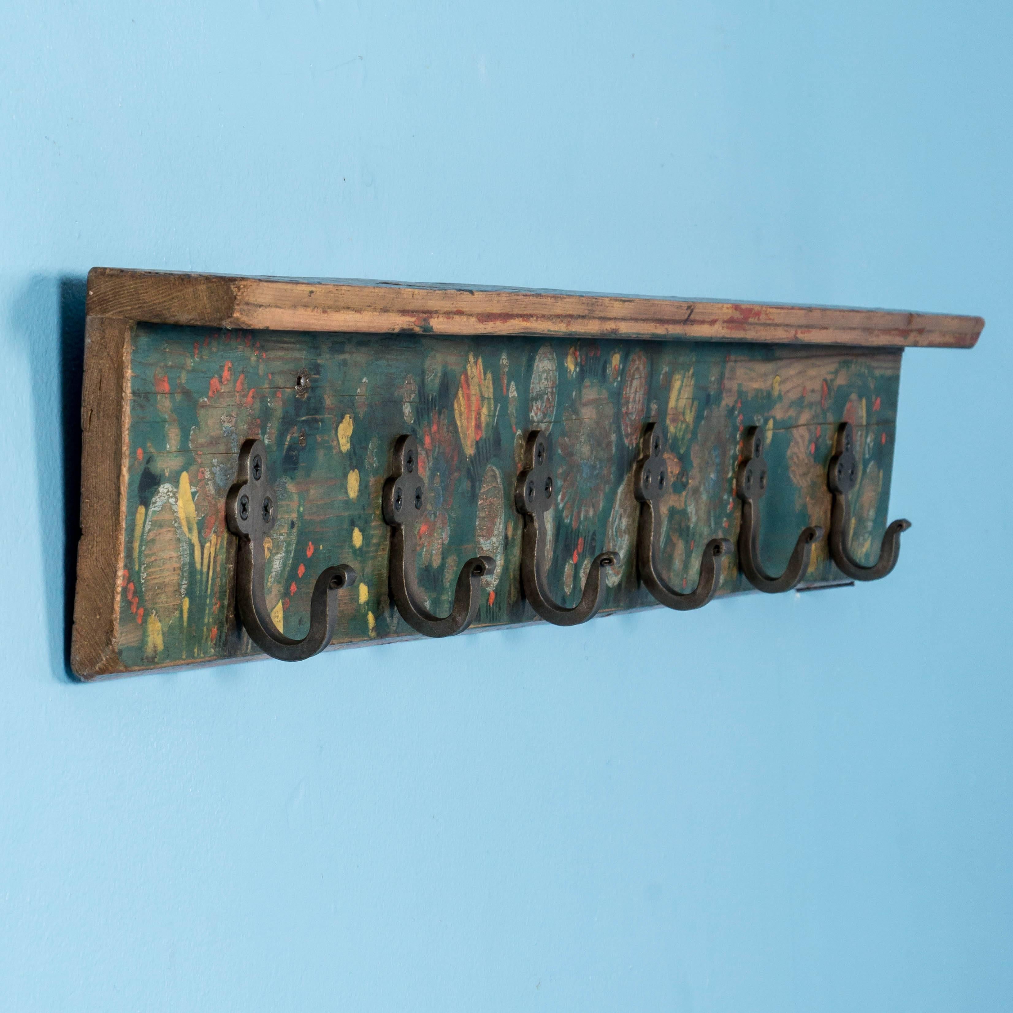 This short hanging wall rack still maintains its vibrant green, yellow and red paint, with Folk Art flowers strewn along the length. While the painted floral design is original, the grey metal hooks are replacements. The painted pine panel has been