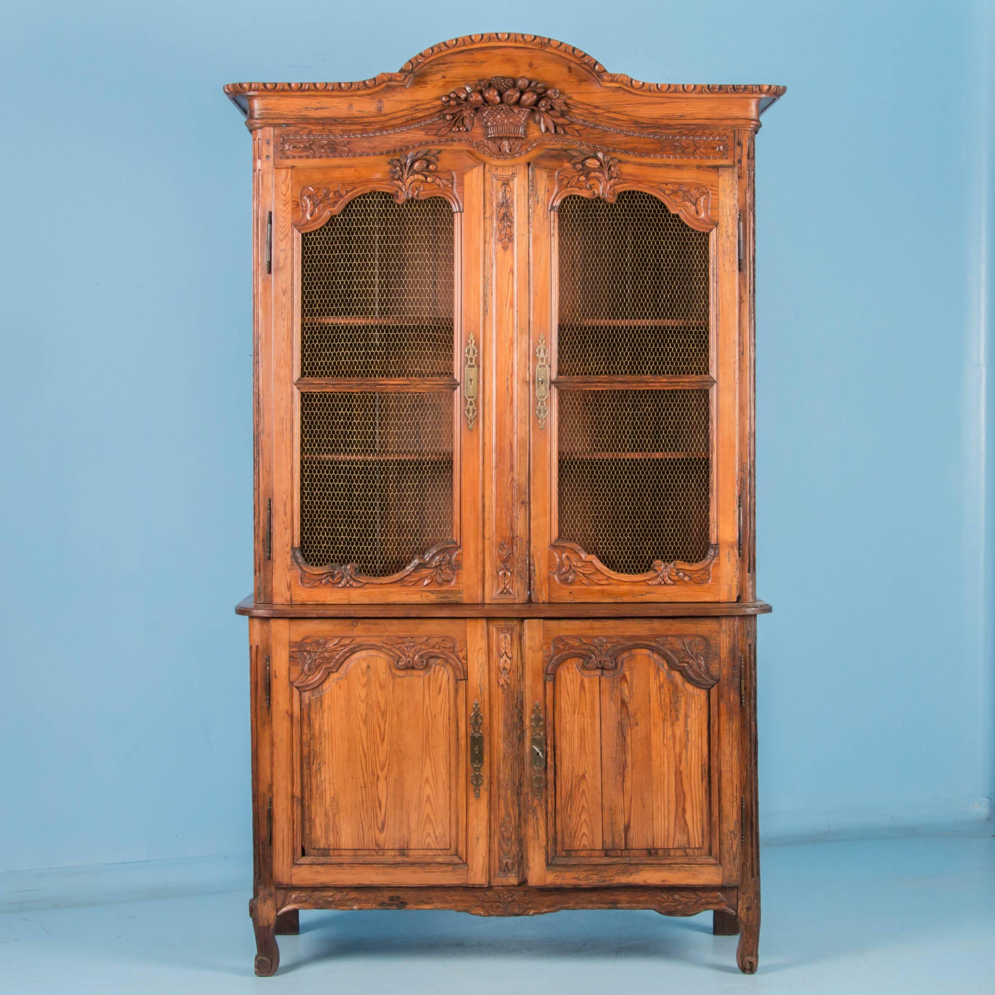 Antique hand-carved four door pine bookcase cabinet from France, circa 1820-1840, made in two sections with the upper doors covered in a honeycomb of brass wire. Elements of the carved woven basket with fruit and flowers in the crown, are repeated