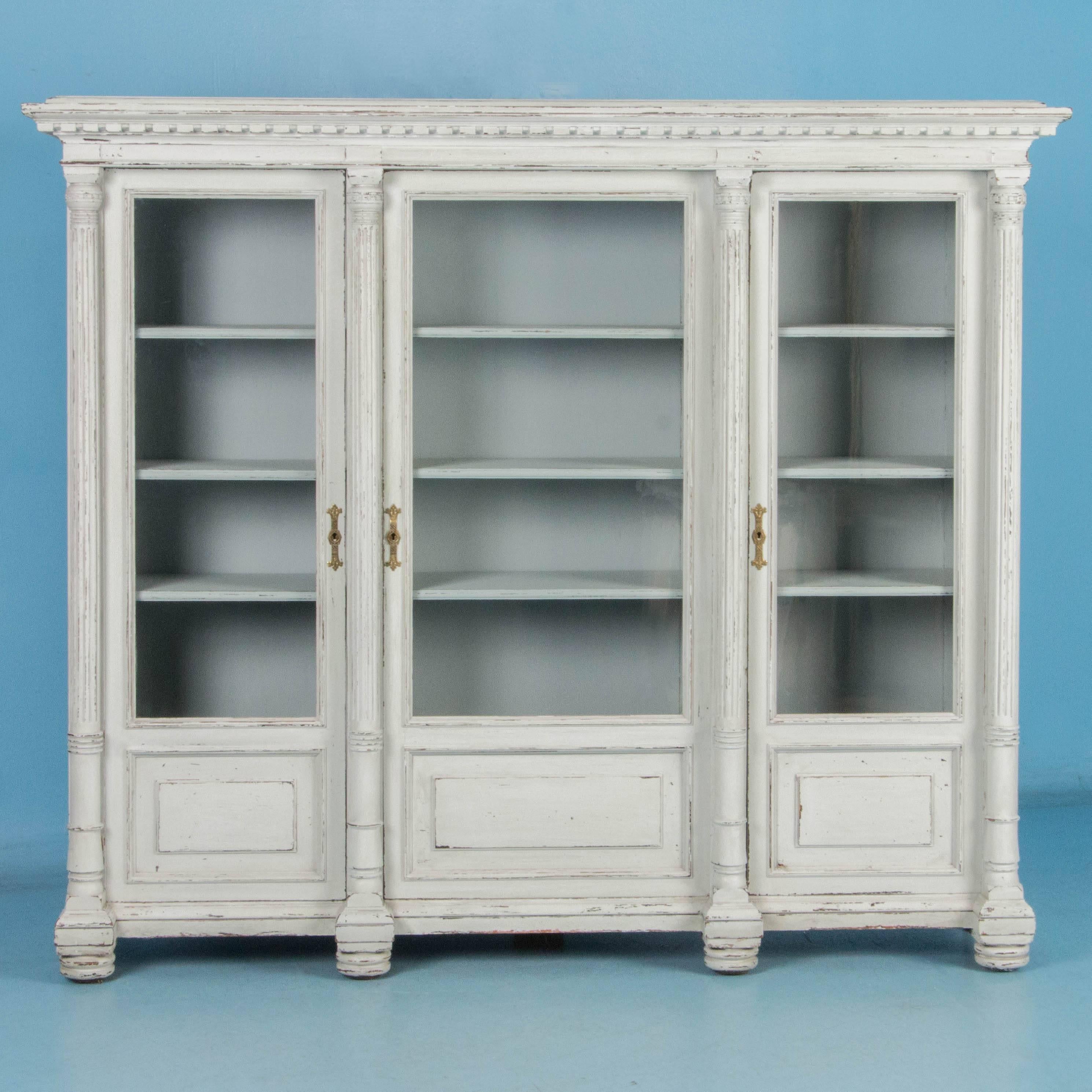 This large antique Swedish oak bookcase (circa 1890s) features dentil molding, fluted columns and panelled doors in a neoclassical design. Behind each of the three hinged glass doors are fully adjustable shelves. The newer white paint had a subtle