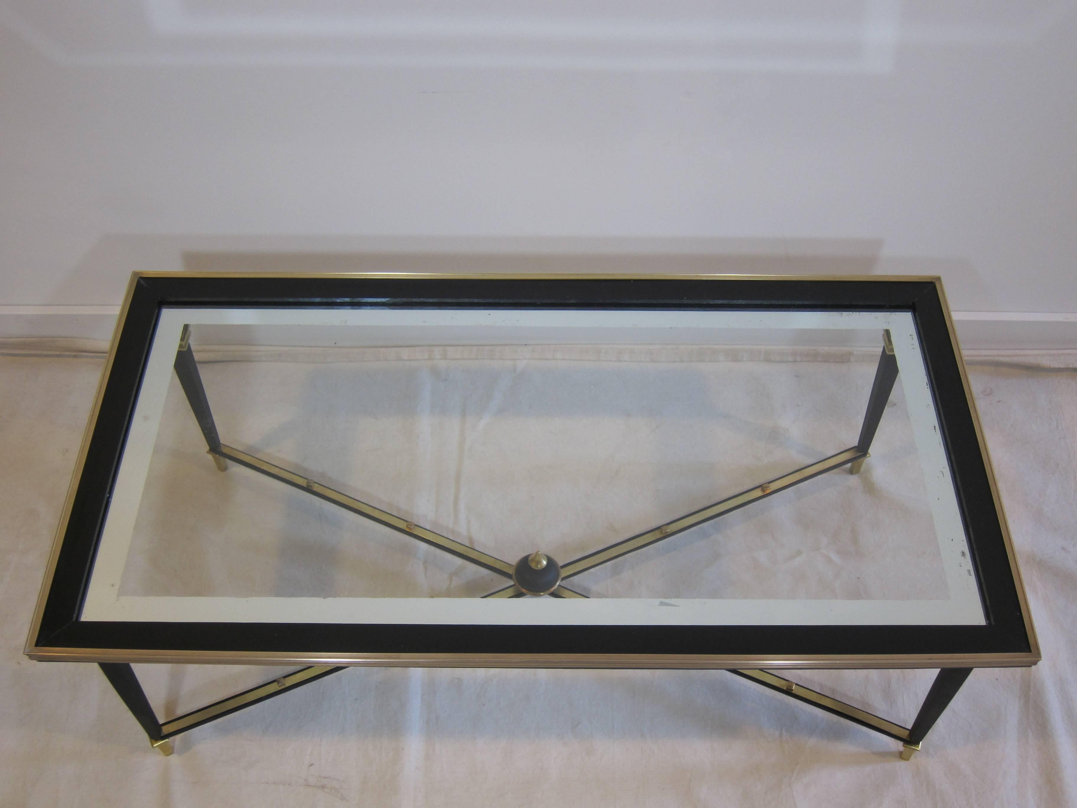 French glass cocktail table in the style of Maison Jansen. Having brass frame with wood frame around a glass top with mirrored border, brass studded struts with brass details. The mirrored silver border has some wear pictured, minimal scratch to the