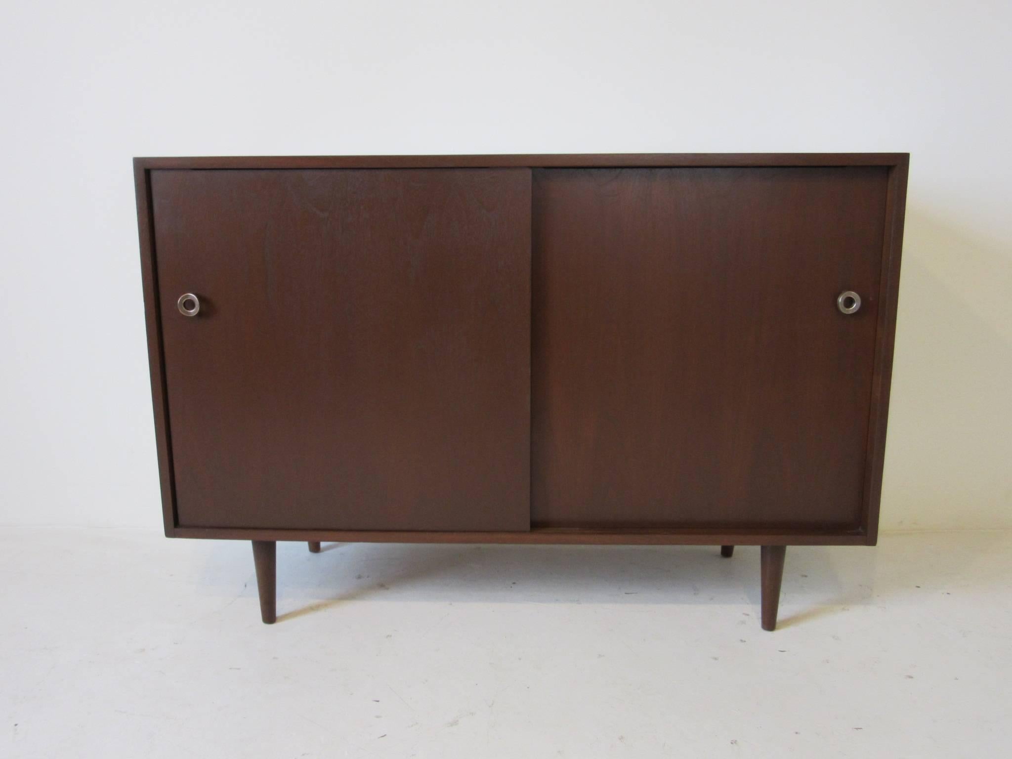 A dark walnut two-door credenza with bronze pulls, storage and adjustable shelve and the other side having four tangerine orange drawers sitting on conical legs. Manufactured by Glenn of California.