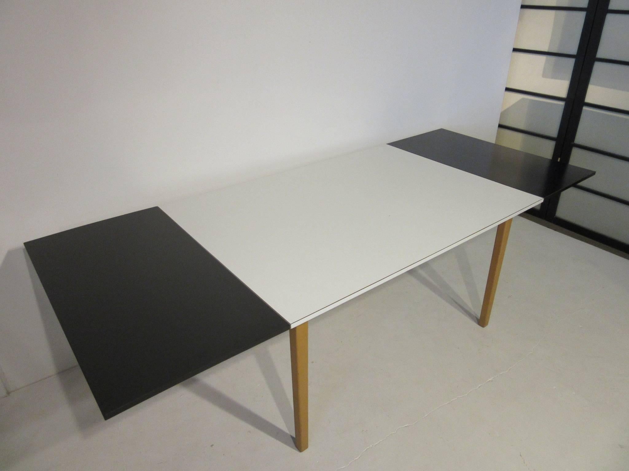 A hard to find and rare Knoll & Drake black and white laminate extension dining table with two black slip in end leaves. The base is a natural colored maple wood with two shallow pull-out drawers to each end for silverware or linens. Designed by