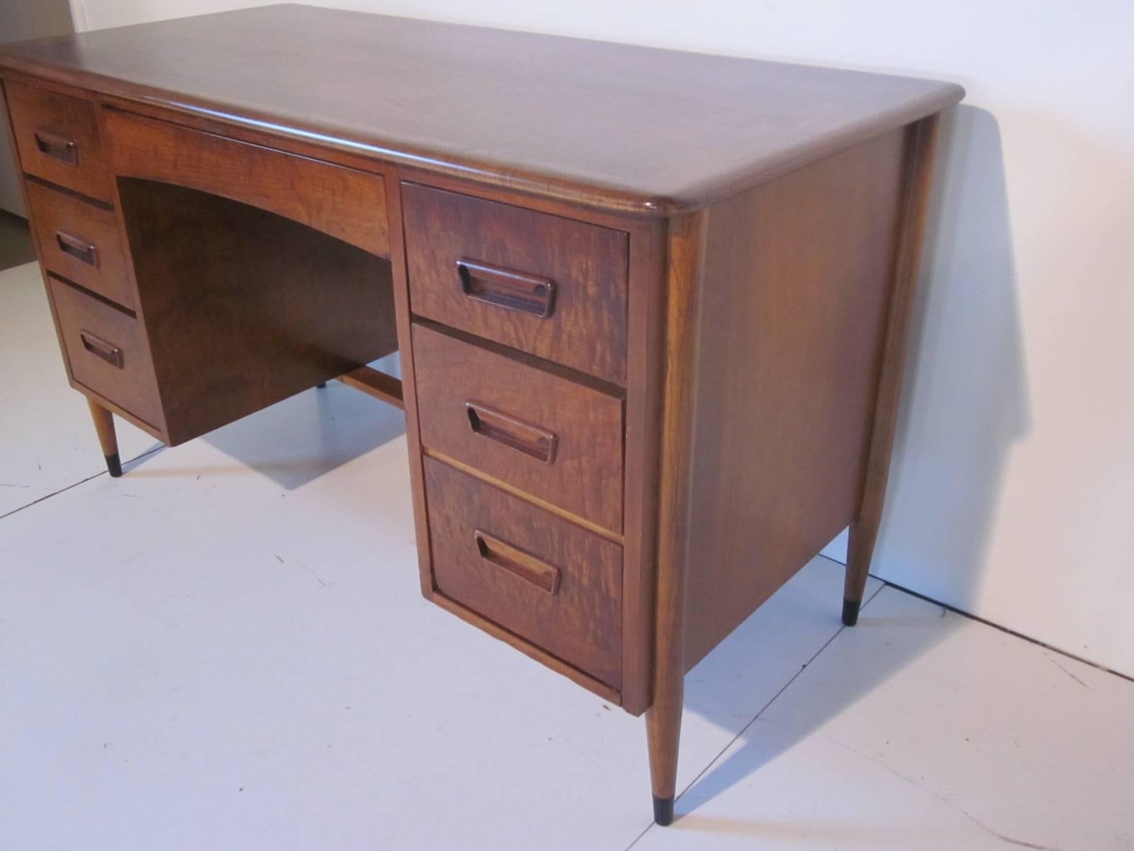 A dark walnut kneehole desk from the Acclaim line featuring lighter dovetail details to each end of the desk top with center pencil drawer, four small drawers and a file. The back side is finished as the front and the conical legs have black end