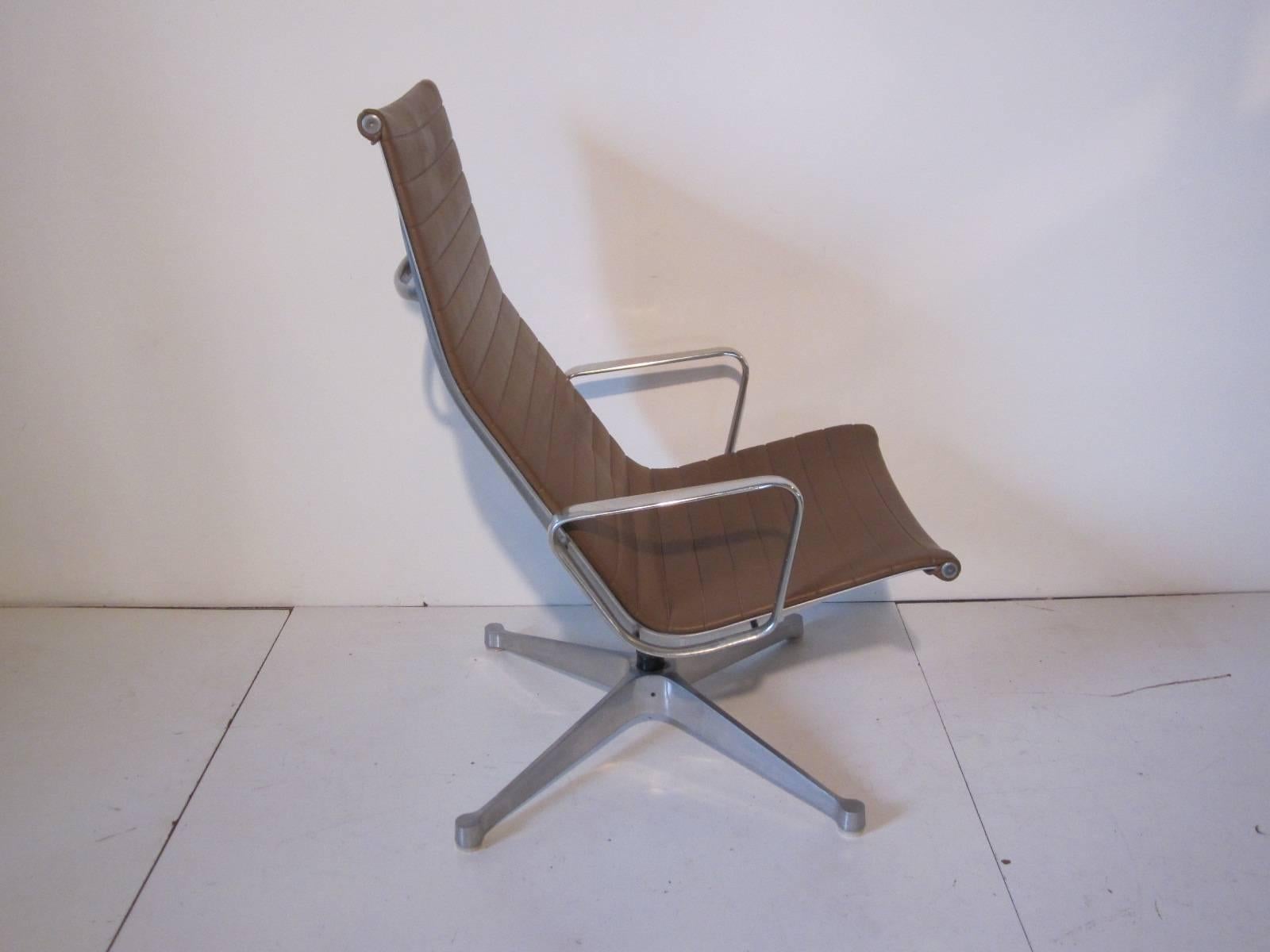 A Eames aluminum group tilting back lounge chair with medium saddle colored Naugahyde seat and aluminum arms and four star base. The chair does not swivel but has the optional spring fitted tilting base. Manufactured by the Herman Miller Furniture