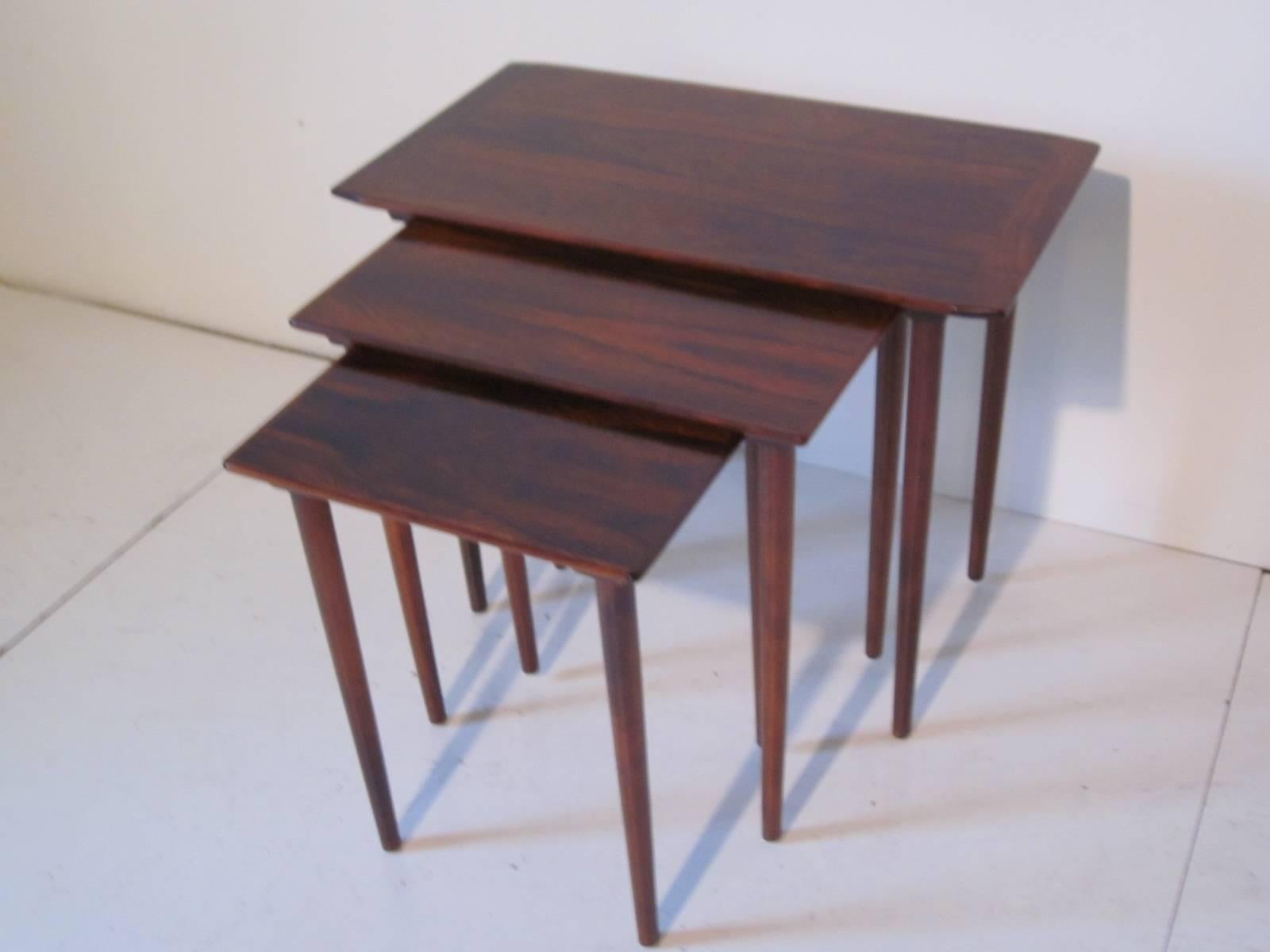 A set of three dark and well grained Danish styled rose wood nesting tables, each slides into a wood grove to secure them until needed. The larger table size is 24.85