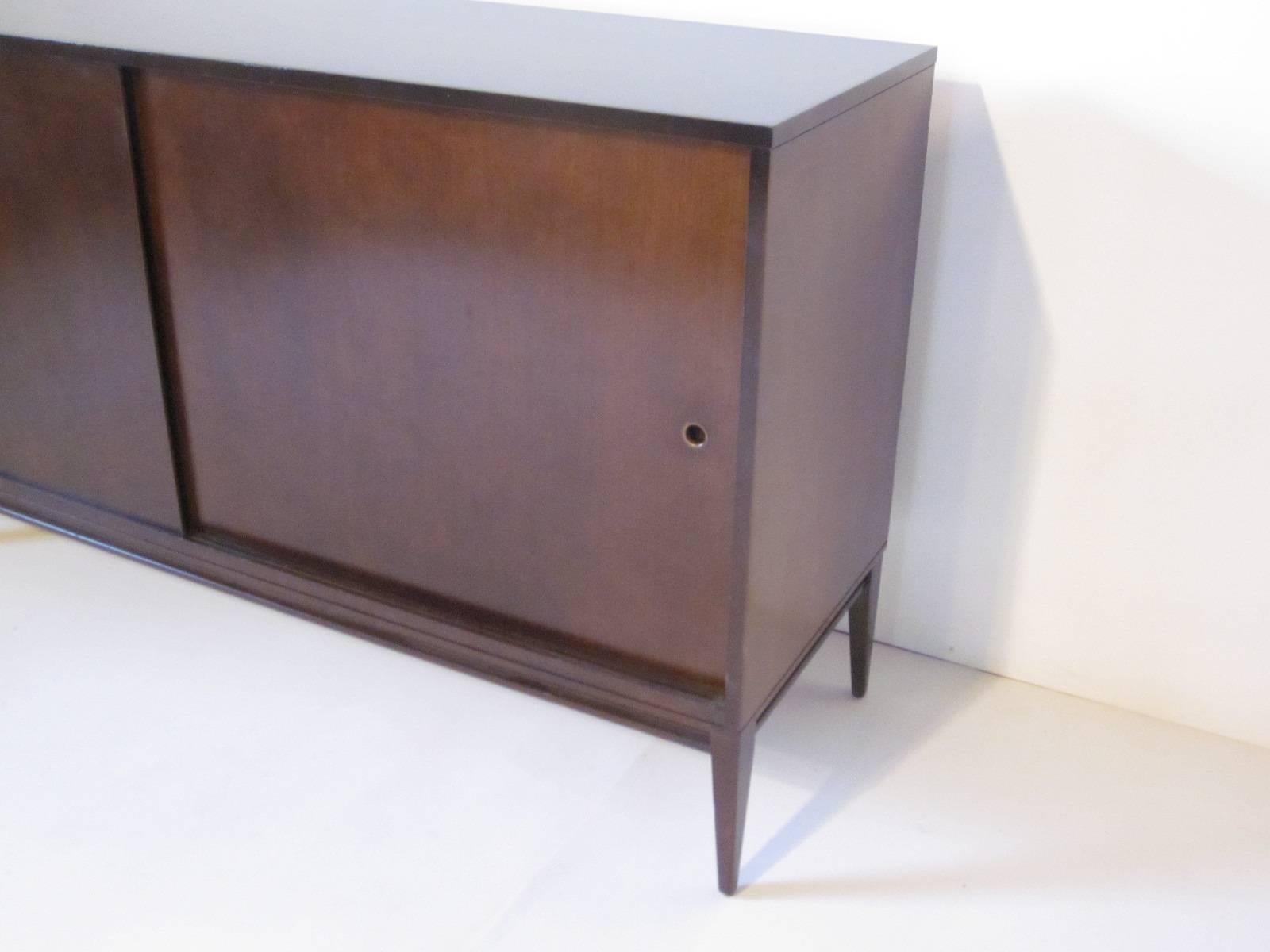 A McCobb dark toned double sliding door credenza with brass hole pulls, four drawers and one adjustable shelve and still retaining it's original foil tag Designed by Paul McCobb for the Winchendon Furniture Company.