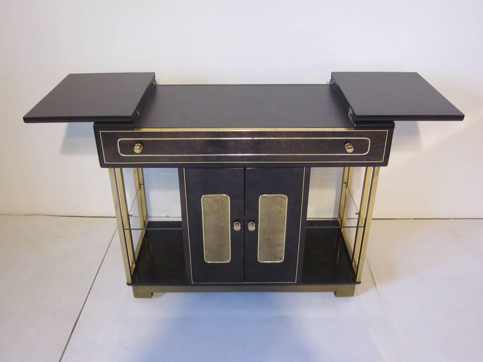 A dark ebony toned burl wood bar or serving cart with acid etched brass door details, internal shelve, brass frame and pulls, a glass shelve to each side, pull-out drawer and wheels. A quality well made piece of furniture with a bit of glam