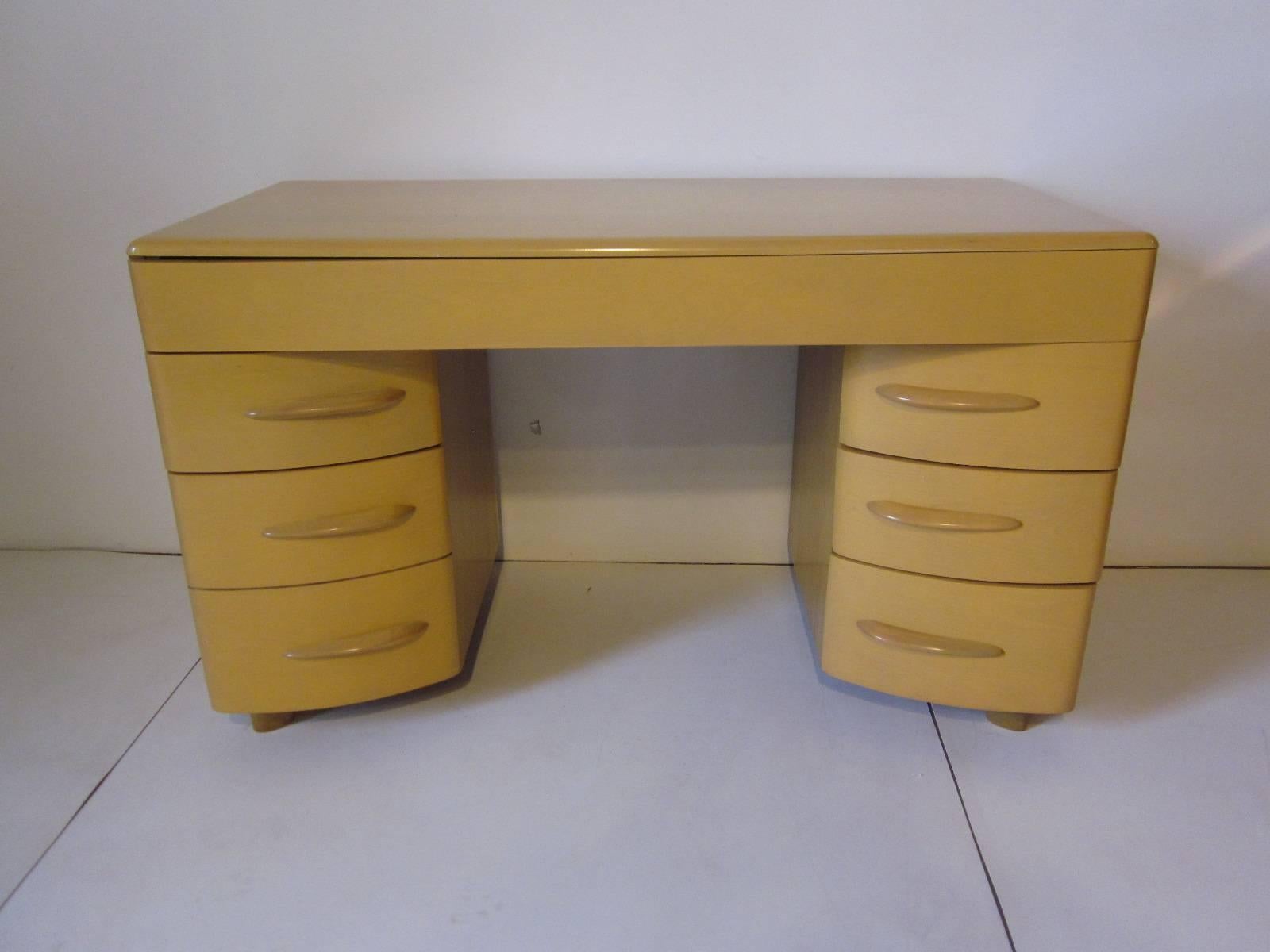 A six-drawer turned in based kneehole desk with upper drawer, one file drawer and four side drawers with streamline pulls. In a wheat toned finish manufactured by the Heywood-Wakefield furniture company.