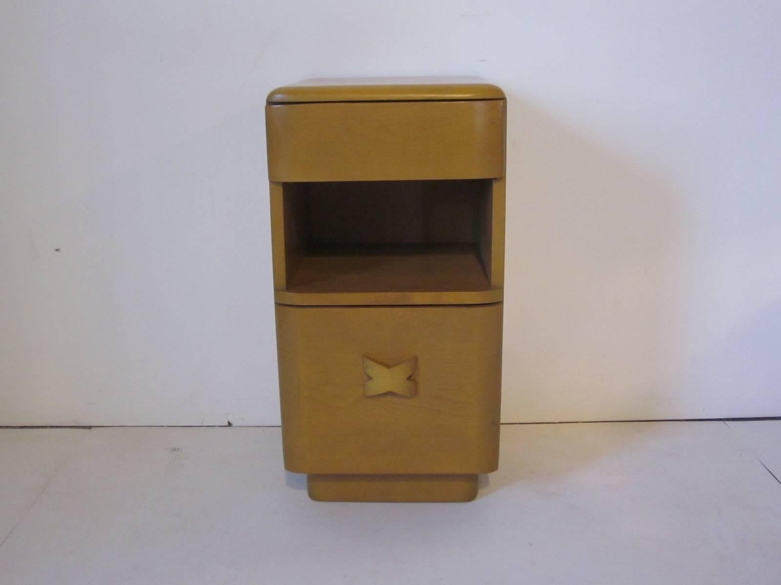 A wheat toned nightstand with single drawer, storage space and lower pull down door with X wood pull. Manufactured by the Heywood Wakefield furniture company.