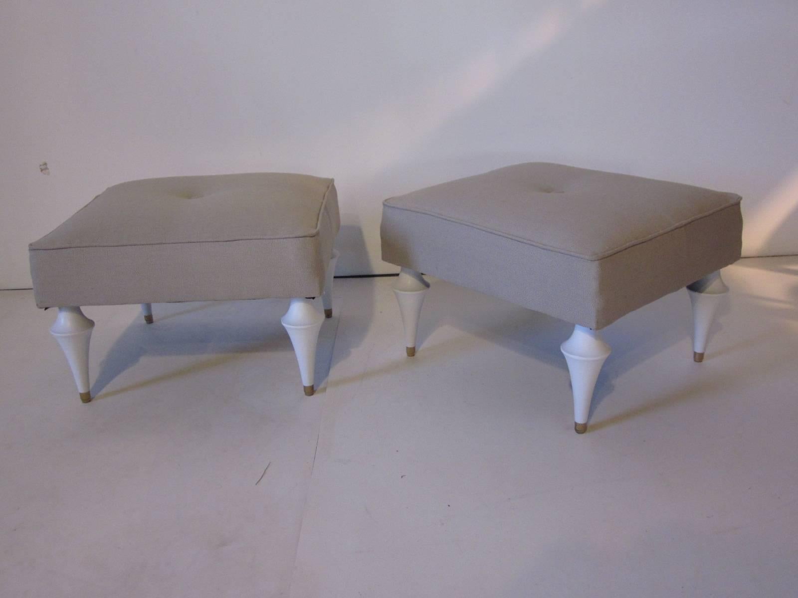 A pair of matching smaller petite sized footstools upholstered in an Italian linen with turned white wood legs tipped in a brass finish in the style of Tommi Parzinger.