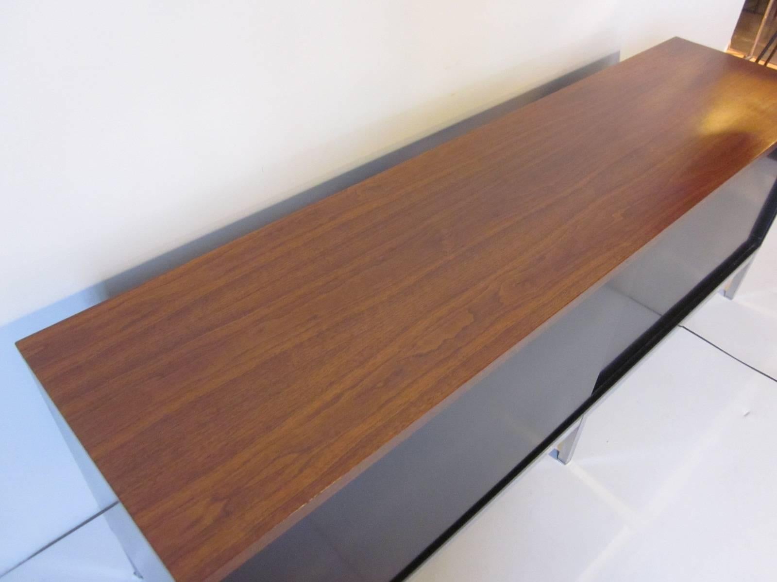 A walnut topped credenza with two sliding doors with aluminum pulls containing four drawers and two adjustable shelves sitting on square chromed bar legs and frame. The body of the credenza is metal and the design attribution is to Florence Knoll