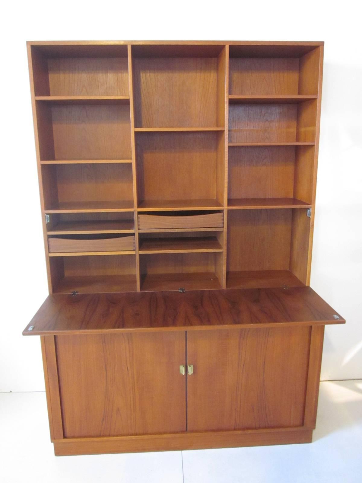 A teakwood secretary and bookcase with lower tambour door credenza, two pull out drawers and shelves with storage. The upper section has a pull down door revealing a desk top with two drawers and shelves. Retains the manufactures markings, ink stamp