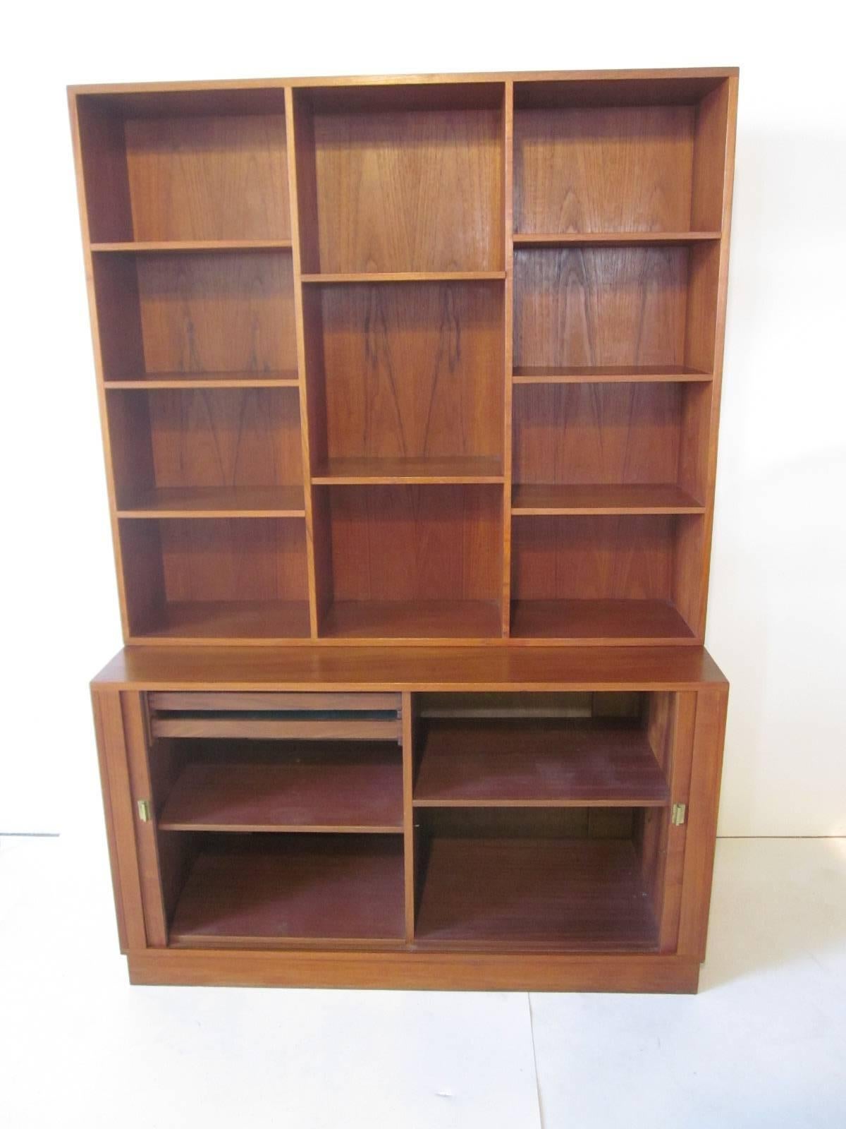 A tambour door two-piece teakwood bookcase and credenza with eight adjustable shelves and lower pull out drawers and adjustable shelves for storage and brass toned metal pulls. Manufactures mark to both pieces with ink stamp date of 1965 and branded