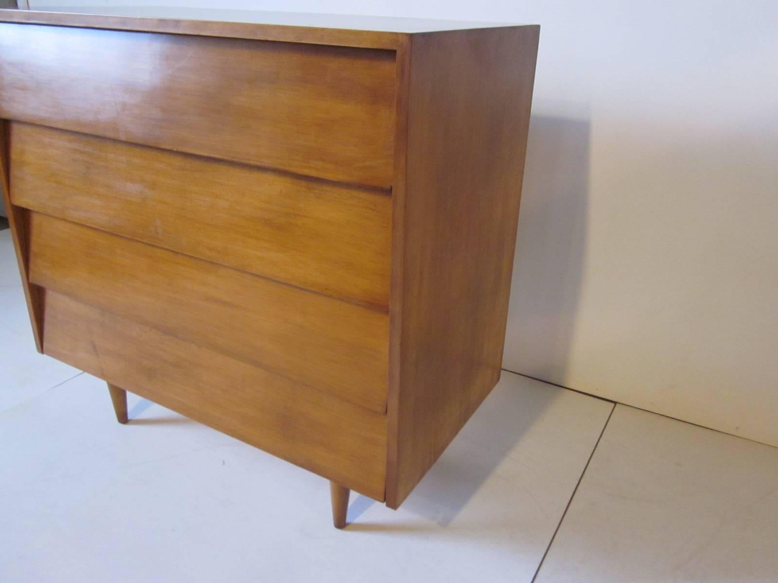 A medium toned maple four-drawer dresser chest with angled drawer fronts sitting on conical legs, a very early design manufactured by Knoll.