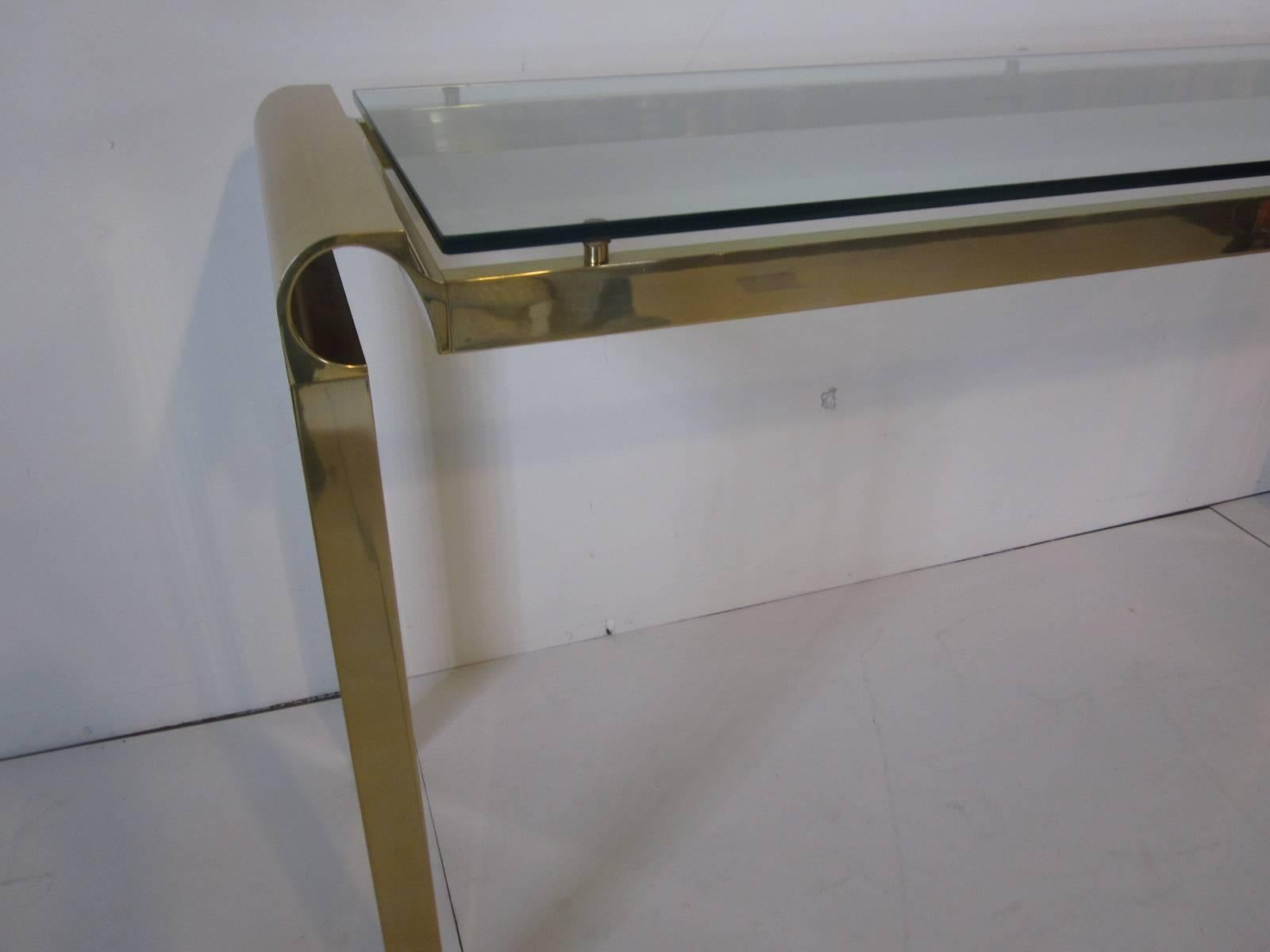 A brass console table in the style of Pierre Cardin with floating plate glass table top and large C-like designs to each corner sitting on square tube legs.