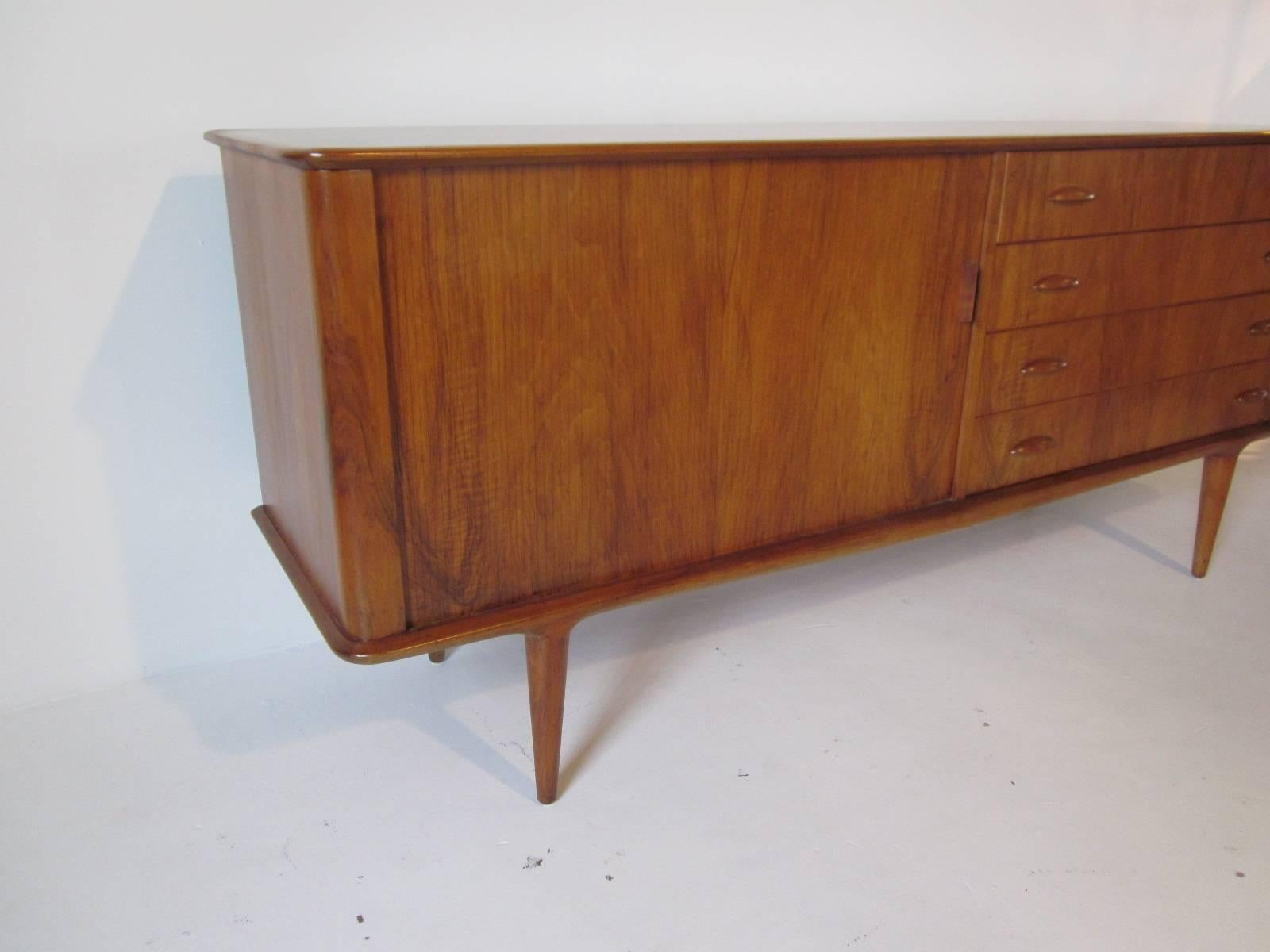 A well grained teakwood credenza or sever with sliding tambour door containing two shelves for storage and a bank of four drawers with wood pulls. Retains the manufactures branded mark M & M Moreddi. Made in Denmark.