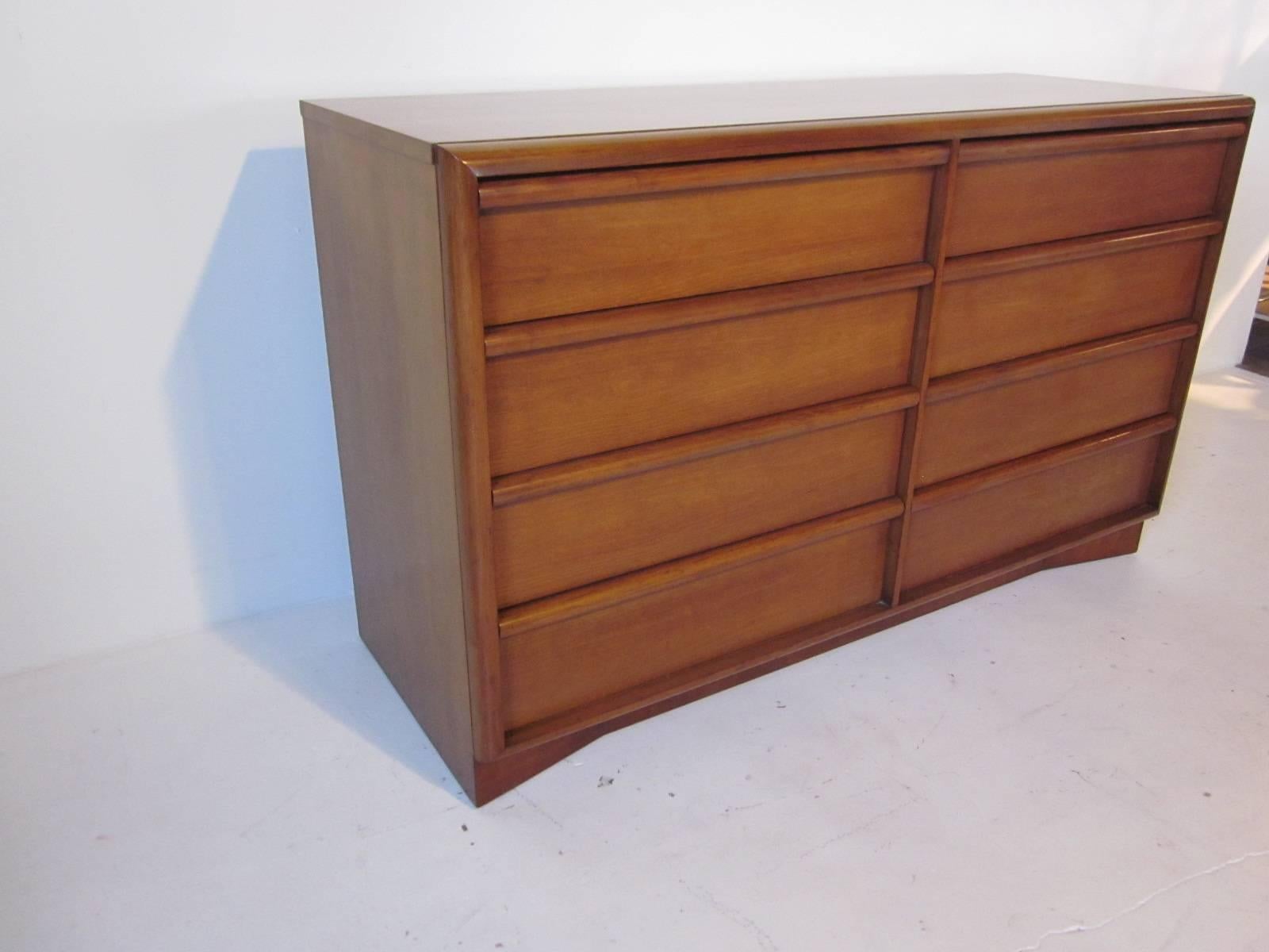 A eight drawer dresser in the manner of T.H. Robsjohn-Gibbings and Widdicomb with a medium dark honey finish true to this style, long pulls to the top of each drawer and curved kick base give it a light feel.