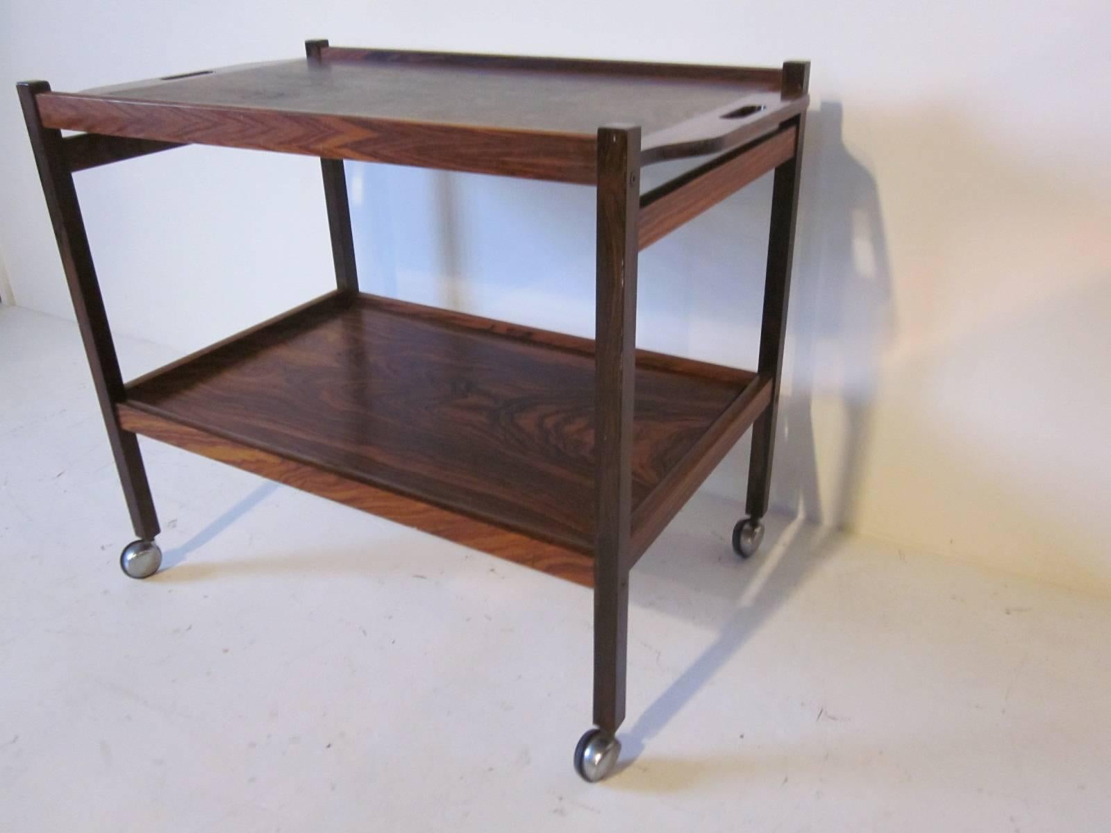 A deep dark Brazilian rosewood serving or bar cart with two shelves and cutout handles to the top edges. Sitting on chrome and black rubber rolling feet, ink stamp to the bottom 