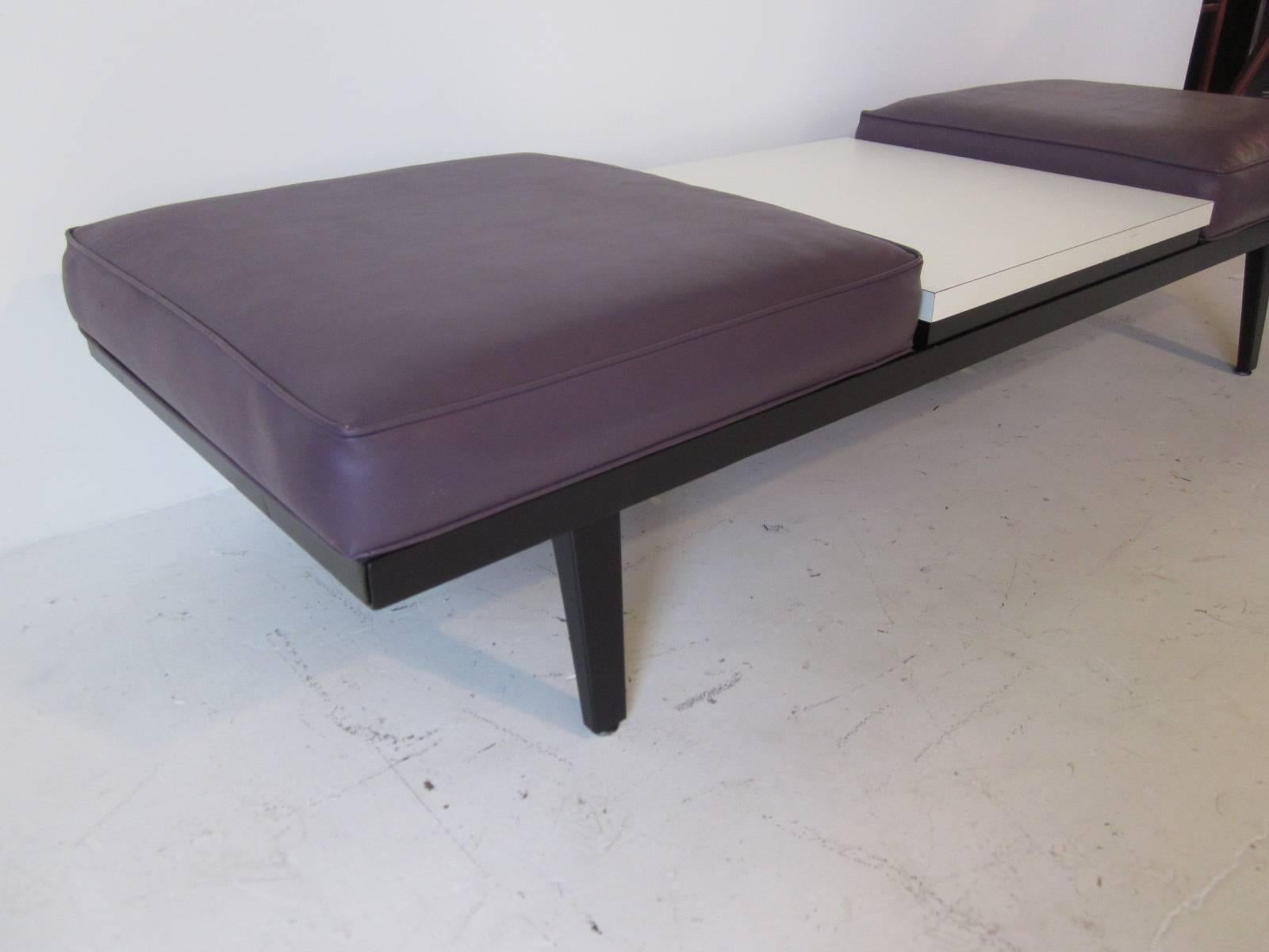 A double cushion Nelson bench with center table in white formica from the steel frame series of furniture by the Herman Miller furniture company. Naugahyde upholstery in a medium purple sitting on a satin black frame complete the bench.