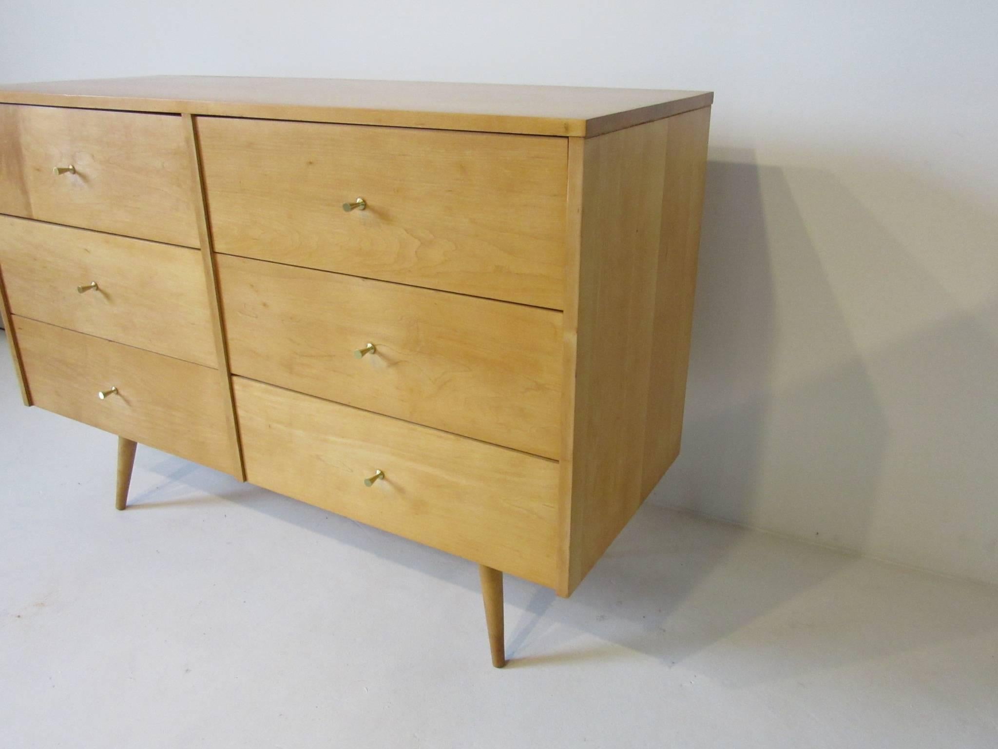 A honey toned solid maple six-drawer dresser chest with brass tee pulls sitting on matching conical legs, from the Planner Group line manufactured by the Winchendon Furniture Company.