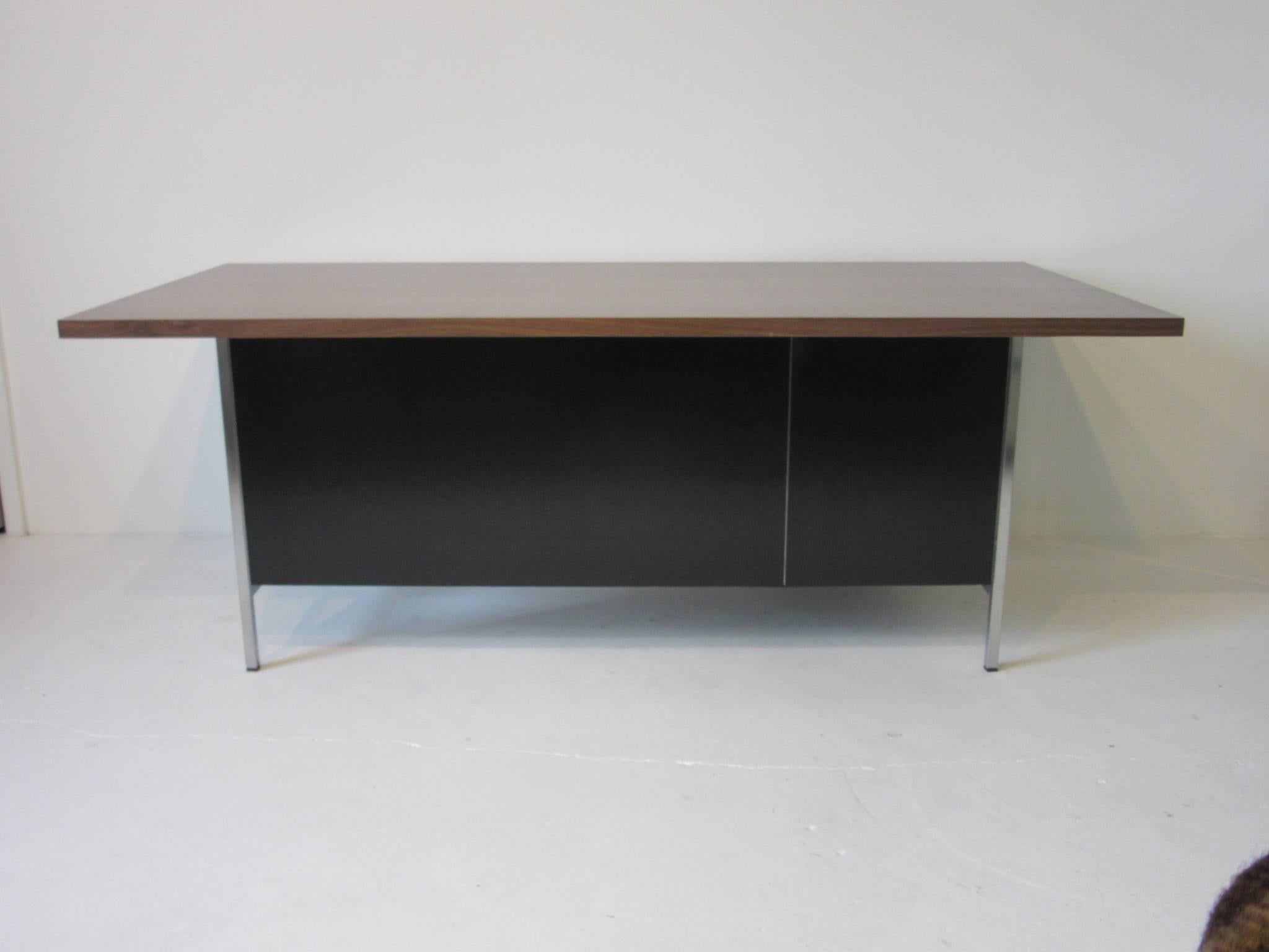 A well designed desk by Florence Knoll with satin black panels and drawer fronts a medium toned walnut desk top all sitting on a brushed stainless steel frame. There are two drawers to the left the lower is a file and the upper one for storage,