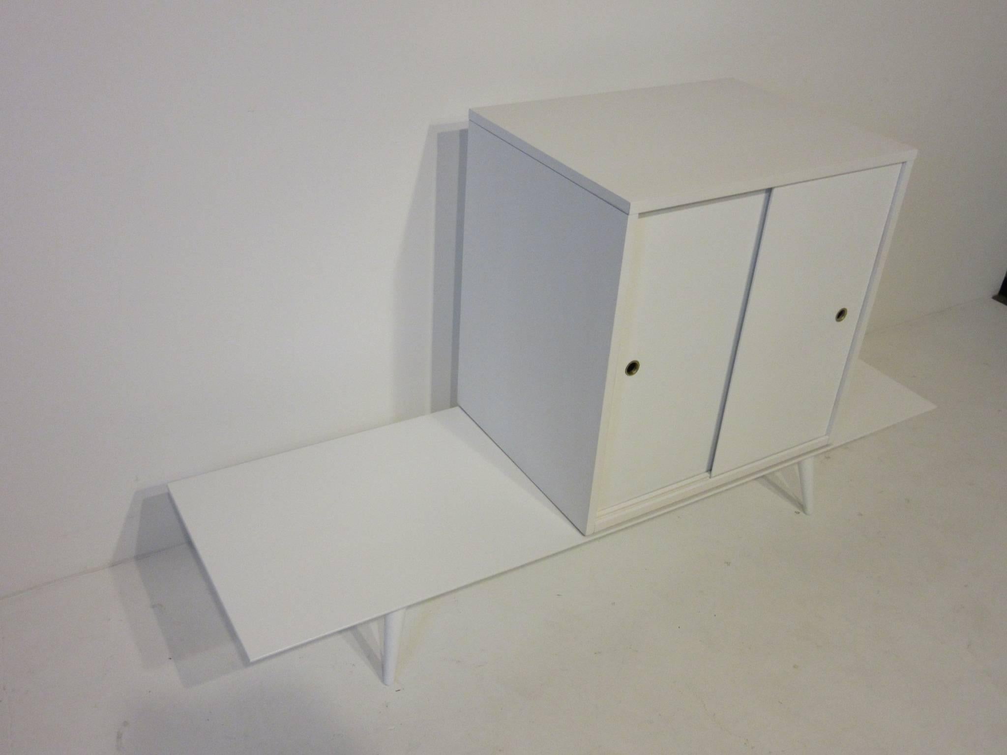 A two-piece Paul McCobb sliding door cabinet on bench with brass pull hole surrounds, conical legs and retaining the manufactures labels. Constructed by the factory in a rare satin white finish which makes this set outstanding. From the Planner