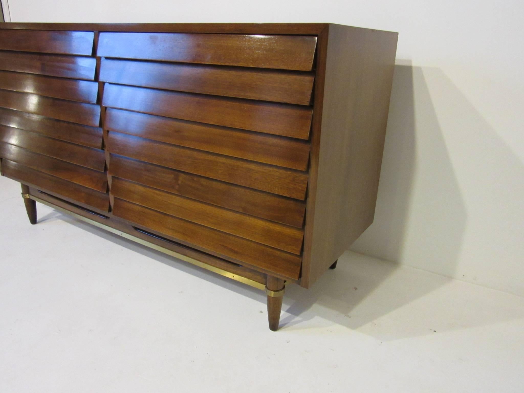 A dark walnut six-drawer dresser chest with louvered front sitting on conical legs with wood and brass detailed stretchers manufactured by the American of Martinsville furniture company.