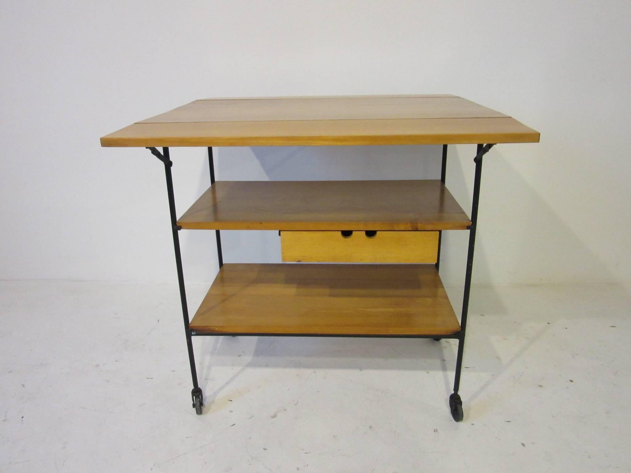 A maple wood bar or serving cart with two folding end pieces making a larger surface a small pull-out drawer on a satin black iron frame and wheels. From the planner group collection for the Winchendon furniture company. Total measurement for the