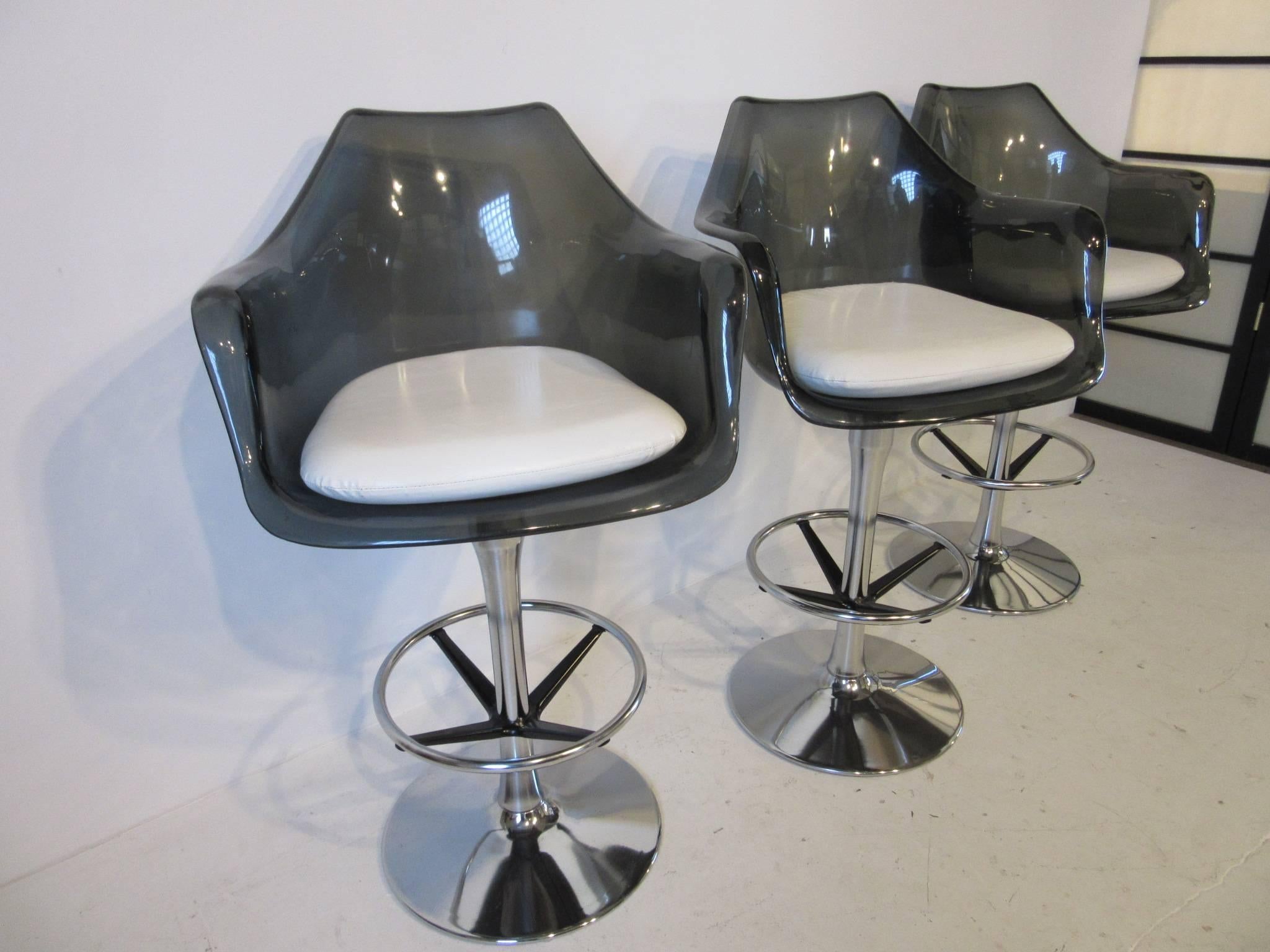 A set of three smoked Lucite bar stools with off-white leatherette upholstered cushions and chrome swiveling bases with foot rests. The seat height is 30