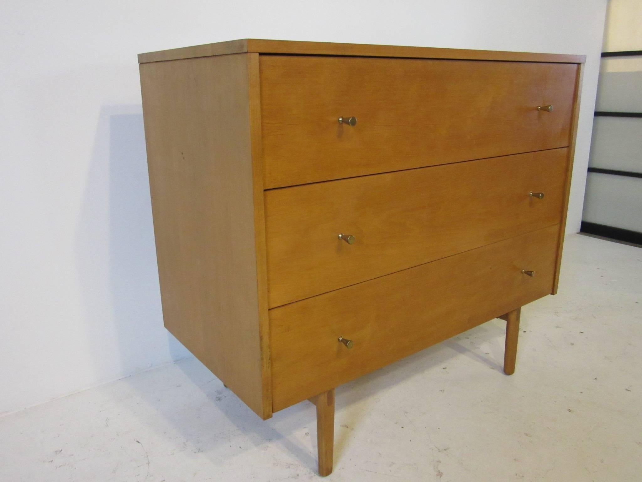 A three drawer dresser chest in maple wood with solid brass golf tee styled pulls sitting on wooden legs. Still retaining the original manufactures label designed by Paul McCobb from the Planner Group Line for the Winchendon Furniture Company.