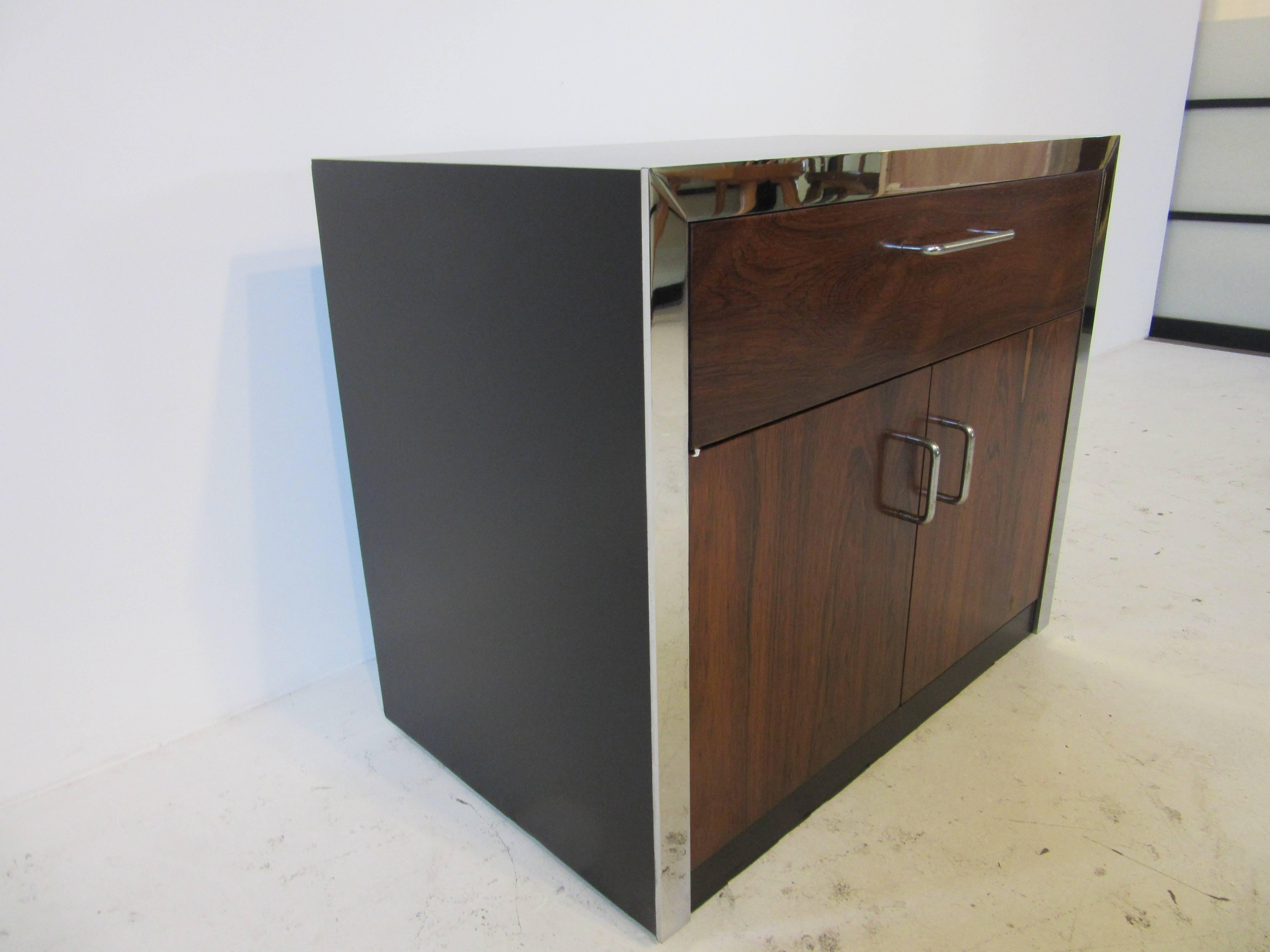 A 1970s Brazilian rosewood nightstand with top drawer and lower double doors with storage. Detail chrome trim surrounds the front with a satin black wood cabinet and chrome pulls. Designed in the style of Milo Baughman.