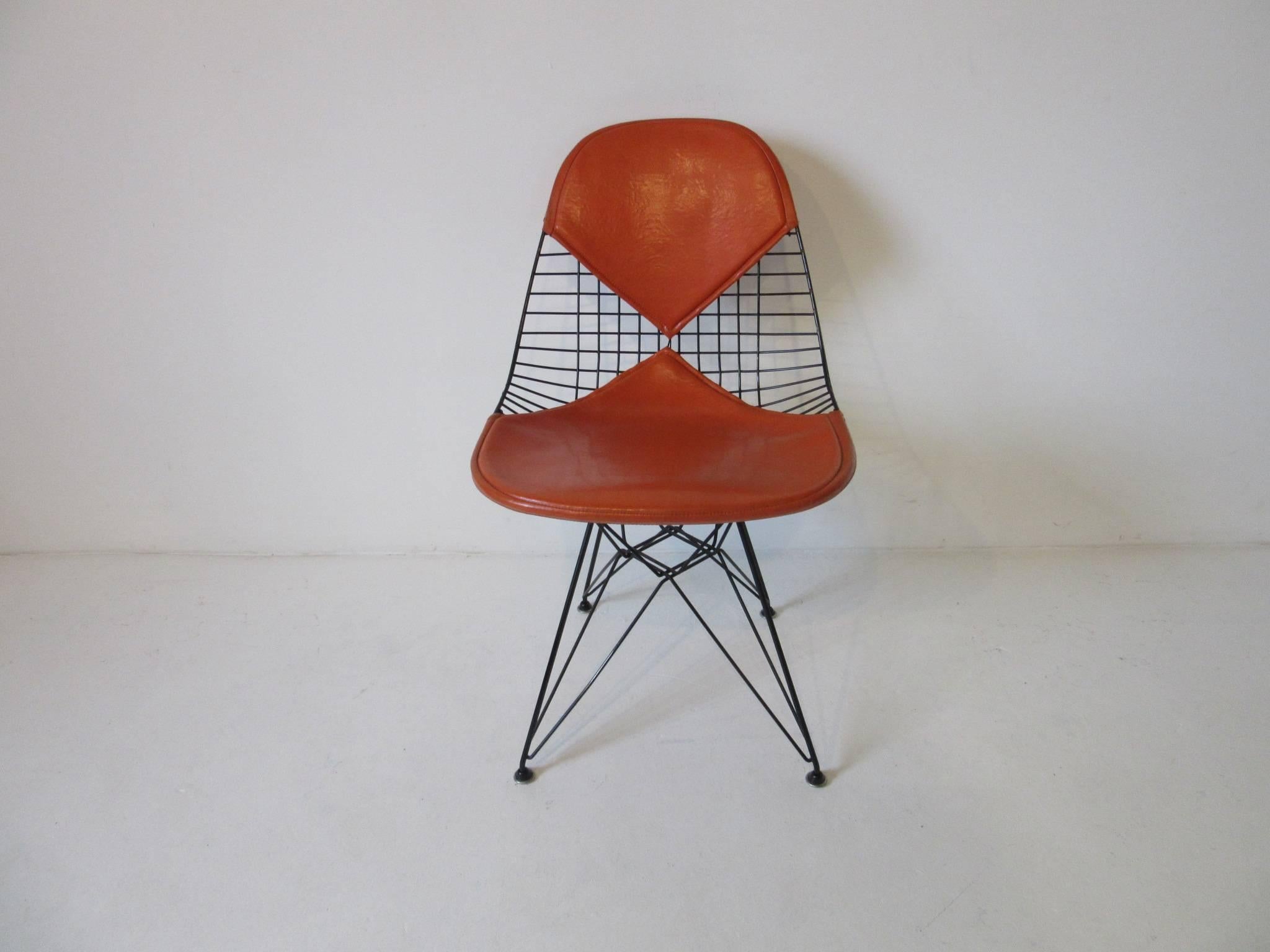 A set of four Eiffel tower based Eames welded wire dining chairs with upholstered orange Naugahyde Bikini seat pads. Earlier production with the self-leveling domed foot pads, manufactured by the Herman Miller furniture company.