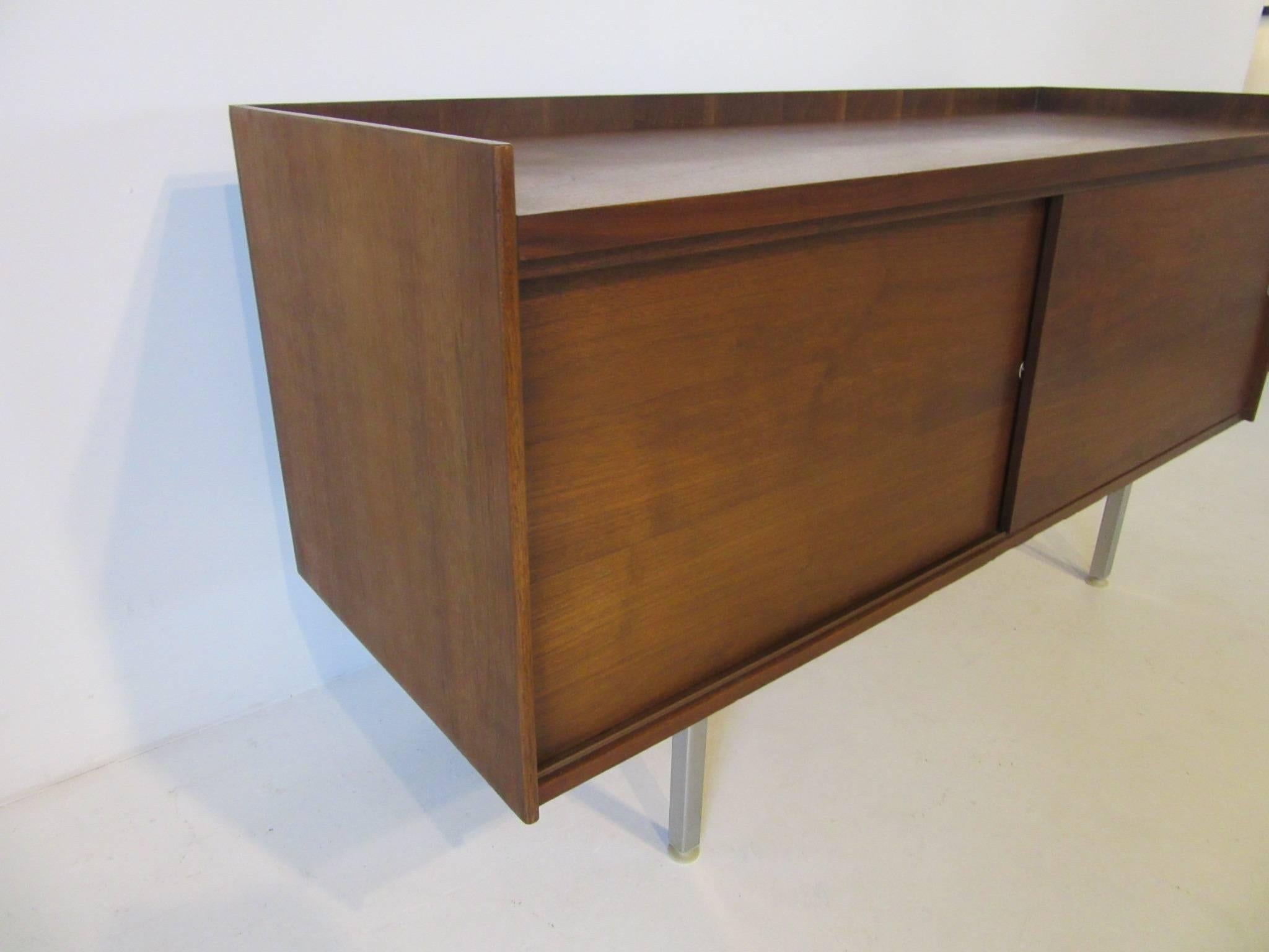 A smaller sized walnut credenza with double sliding doors, an adjustable shelve to each side and raised top edge all sitting on stainless legs. Designed in the style of George Nelson and Jens Risom.