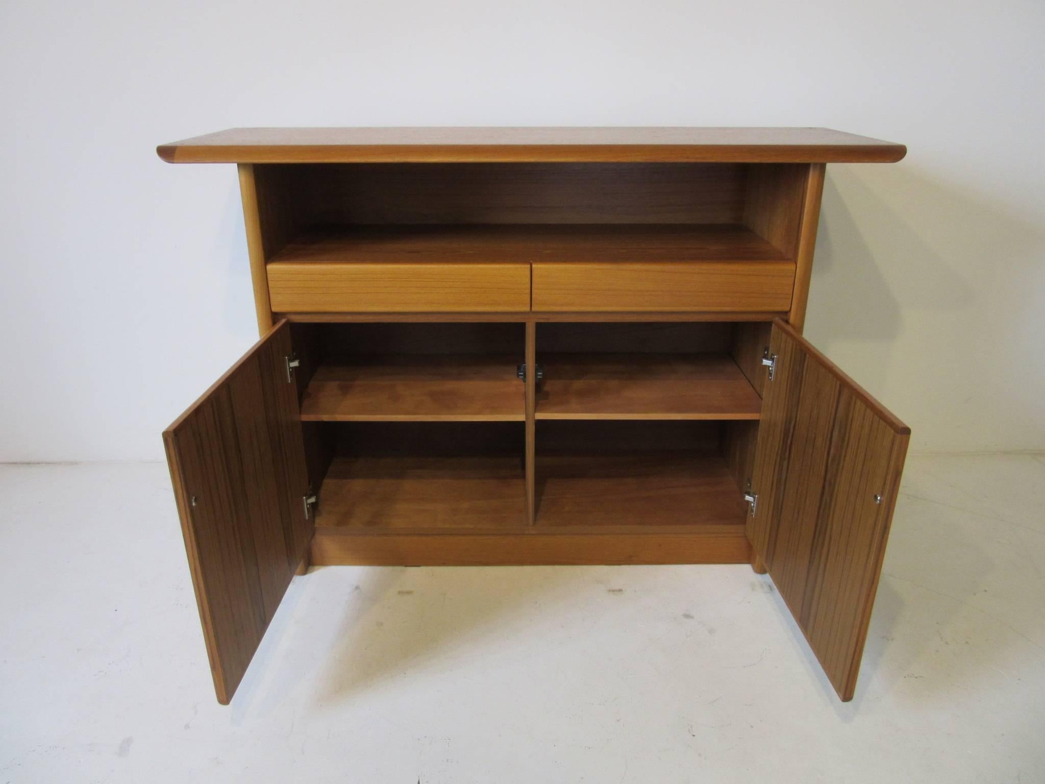 A medium toned teak wood server or bar with open storage and two upper drawers and two door lower storage area with adjustable shelves. Smaller in size and perfect for a tighter space or entertainment area.