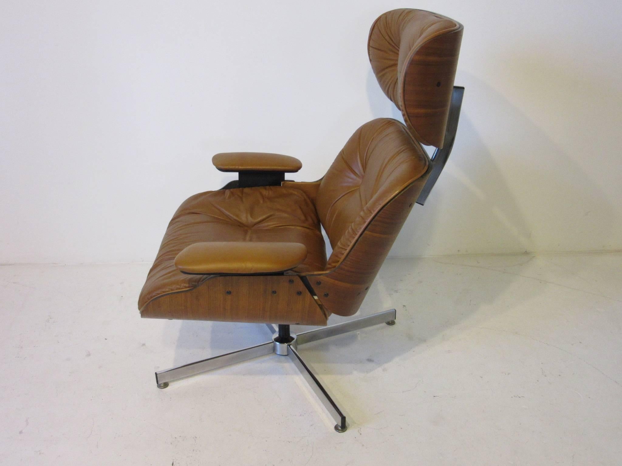 A Mulhauser designed lounge chair in cognac leather with walnut body and chromed metal base. The Plycraft model has an adjustable spring to the bottom for rocking and rake, unlike the Eames model which has a set angle and is uncomfortable to some.