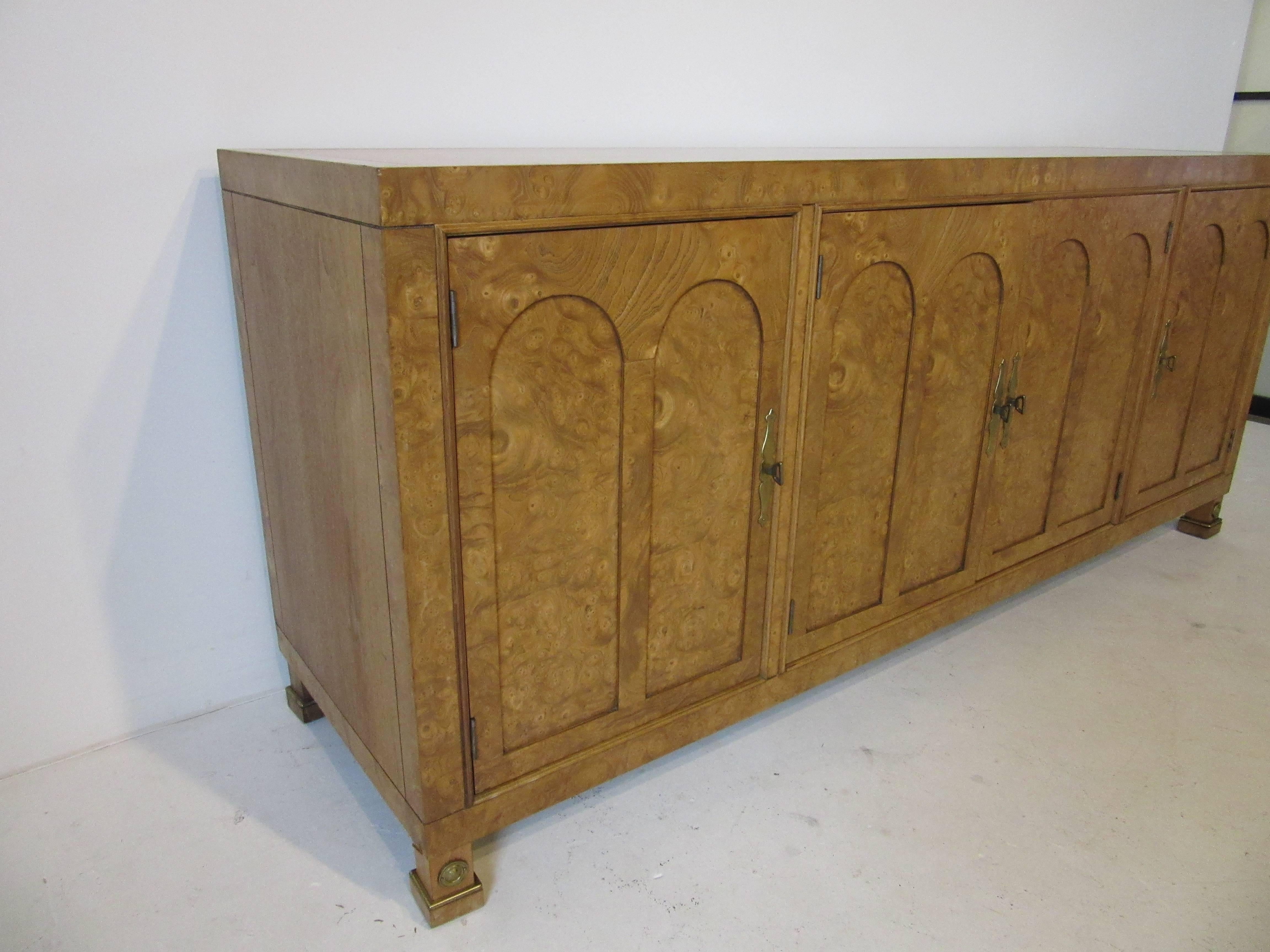 A four door burl wood credenza or sever with brass pulls and details, bookmatched front, columned legs, three drawers and two storage areas with an adjustable shelve to each end. Manufactured by the Mastercraft Furniture Company.
