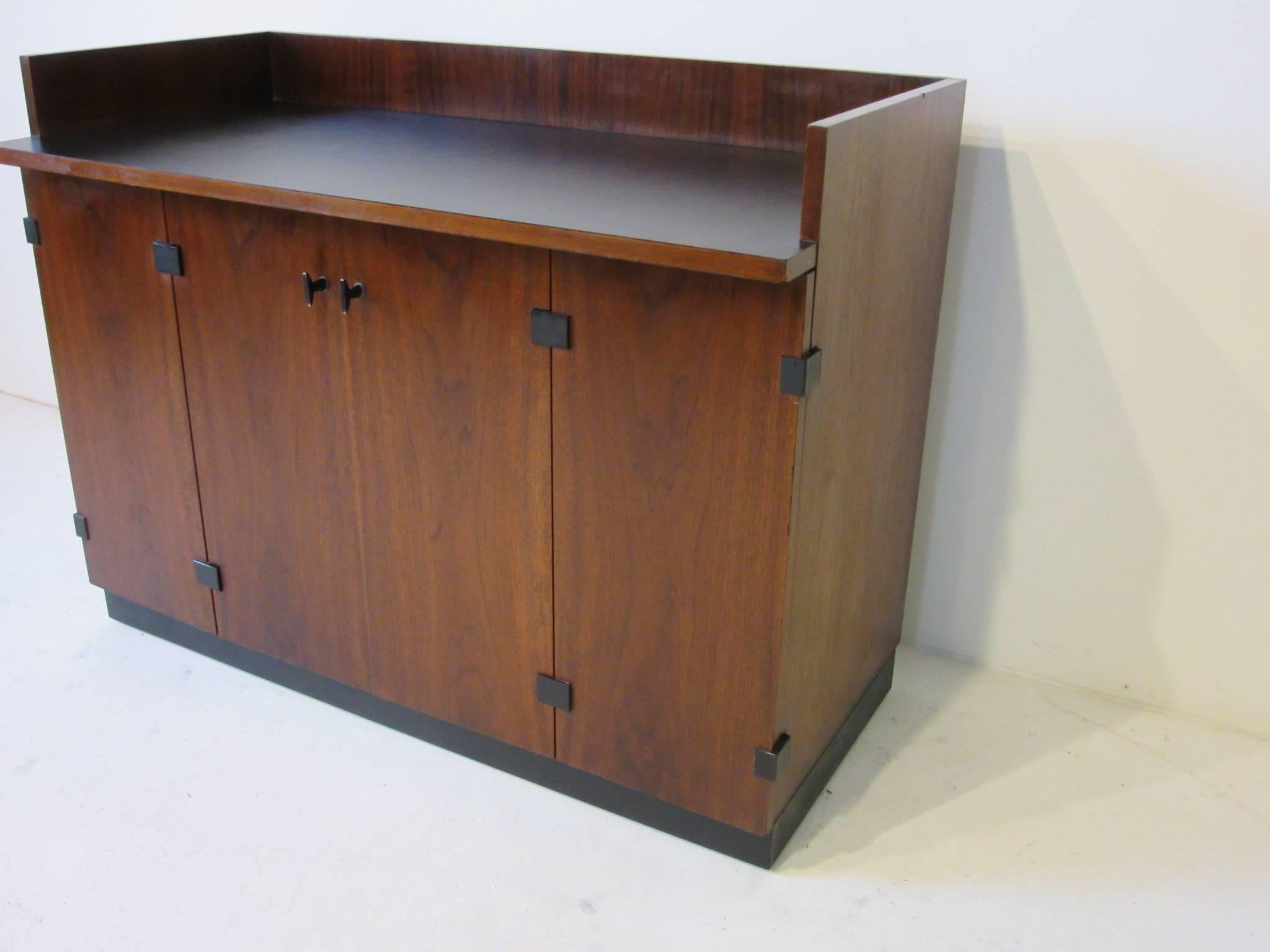 A dark walnut rolling bar cabinet with bi-folding doors, satin black Formica work top, adjustable storage shelve with cut-out for larger bottles, small drawer for bar tools and black detailed hardware. Can be used as a bar or server, finished in