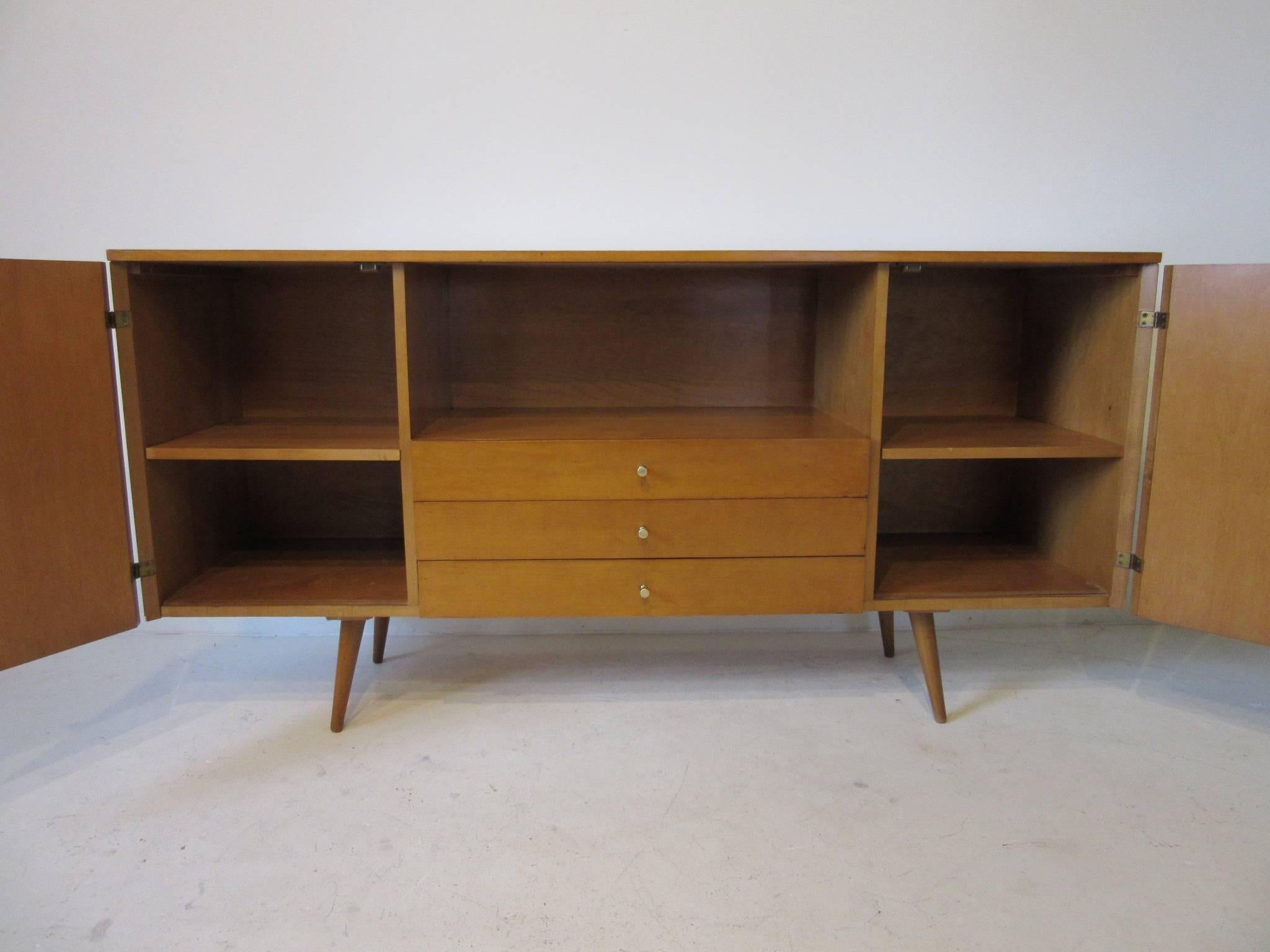 A McCobb credenza or server in a solid maple wood with two doors, adjustable shelves, three middle drawers , brass pulls and an open storage area all sitting on conical legs. A hard form to find which could be used as a entertainment unit, server or