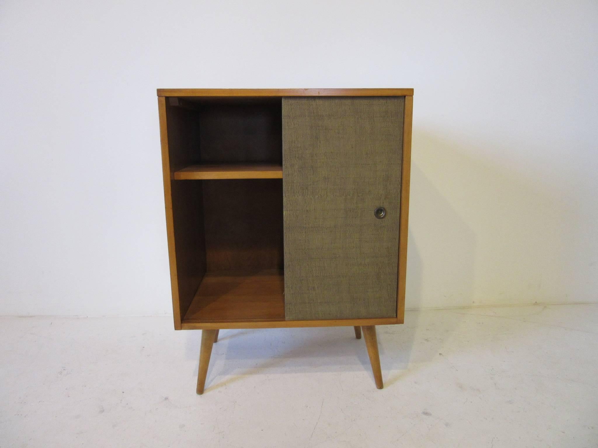 A smaller sized two piece McCobb solid maple wood credenza storage unit sitting on a smaller platform with conical legs and having two sliding grass cloth doors, manufactured by the Winchendon Furniture Company from their Planner Group Collection .