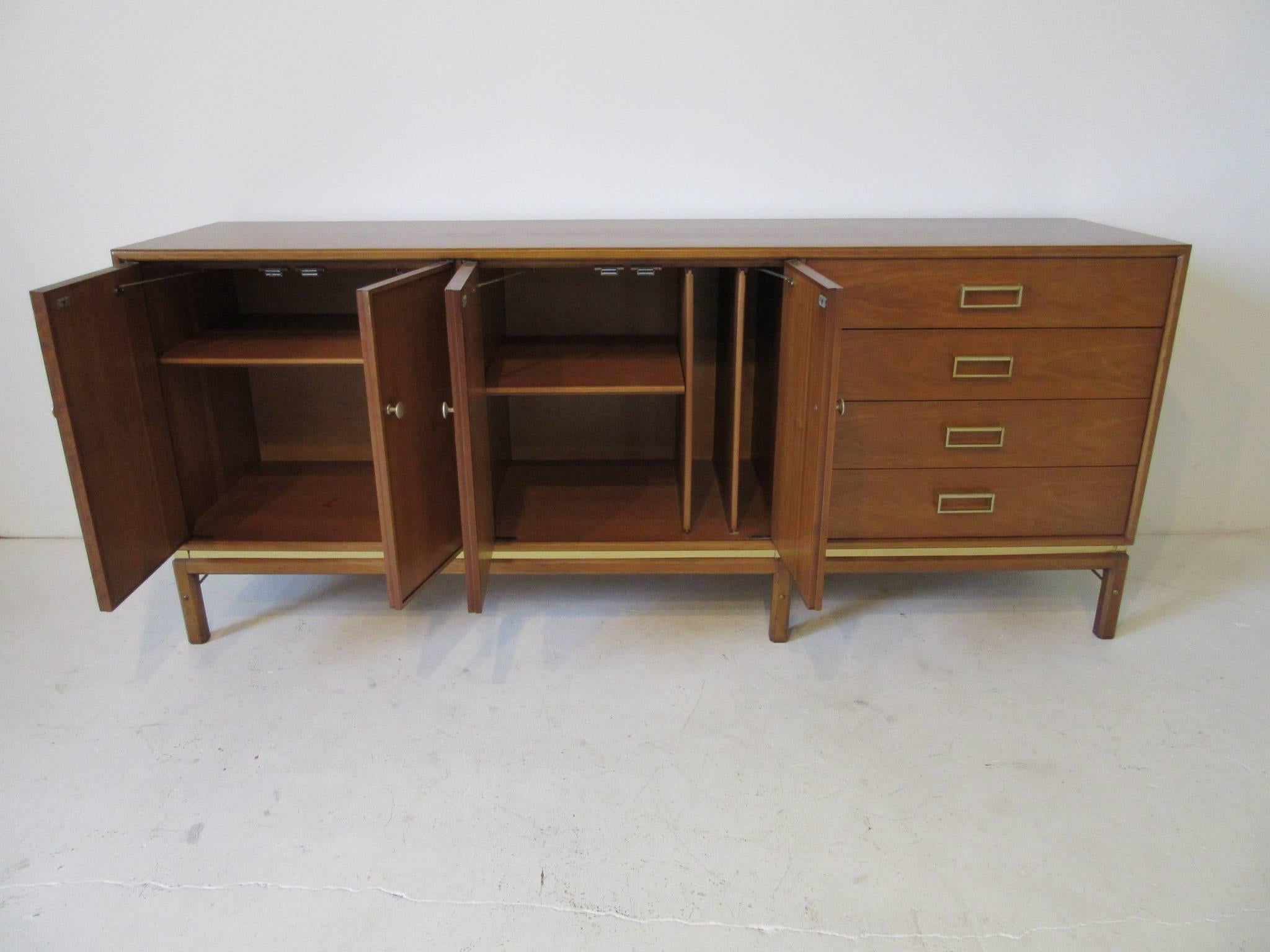 A medium toned mahogany server credenza with brass pulls, lower stretchers and brass details, with double doors, two adjustable shelves and slotted tray or platter storage area. Four drawers with hinged handles are to the right that provide ample