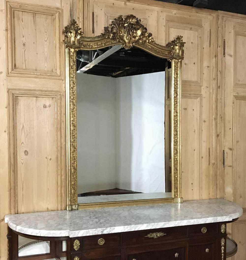 This elegant 19th century French Napoleon III gilded mirror is the essence of opulence with bold crest, floral and foliate adornment on the gracefully arched crown flanked by urns bursting with more florals and a lavish foliate pattern surrounding